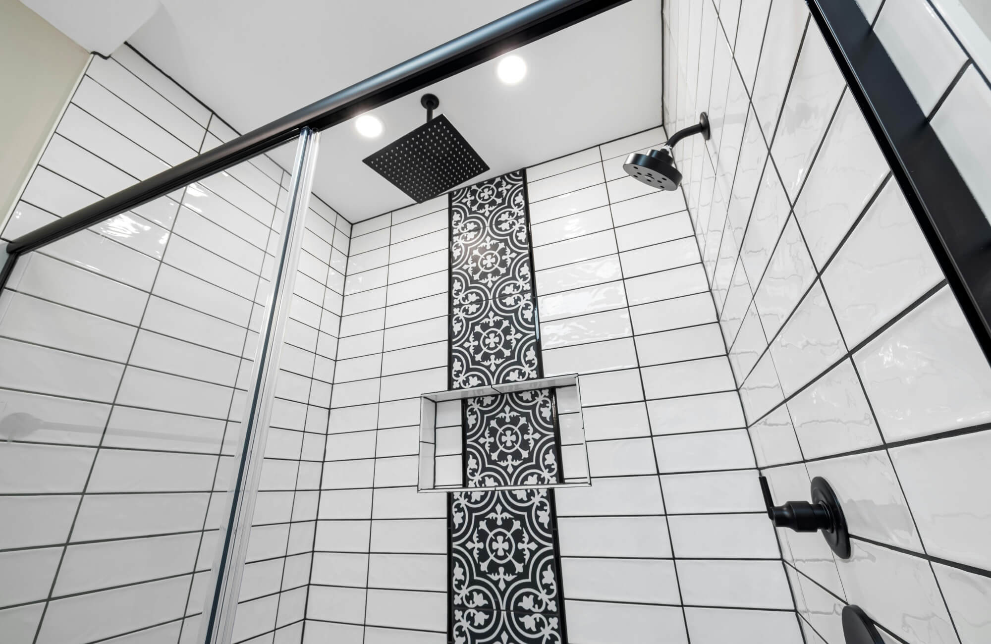 Walk-in shower with glossy white subway tiles and black grout, featuring patterned black accent tile strip and matte black shower fixtures.