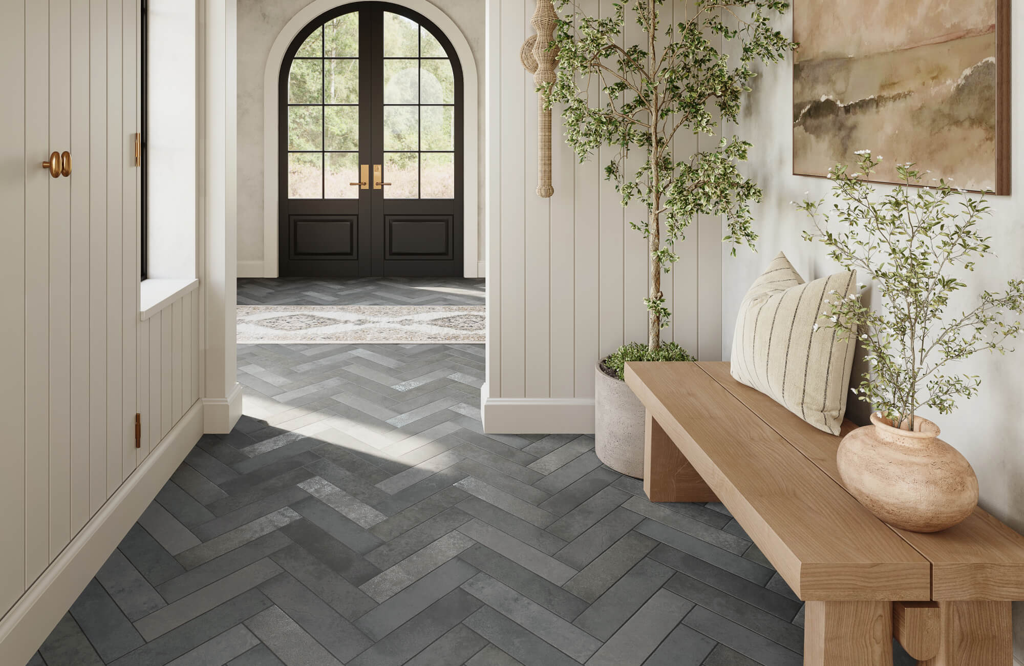 Light-filled entryway with a herringbone zellige-style tile floor, wood bench with pillows, potted greenery, paneled walls, and an arched black front door.