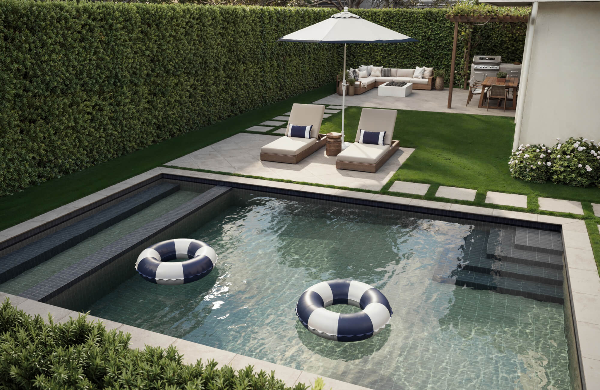 Modern backyard with a rectangular pool finished in zellige-style tiles, floating swim rings, stone steps, sun loungers under an umbrella, manicured lawn, and hedge-lined privacy.