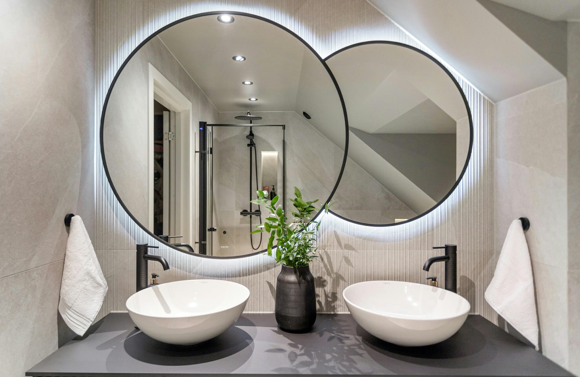 Modern double vanity with matte black fixtures, vessel sinks, and overlapping backlit round mirrors beneath a sloped ceiling.