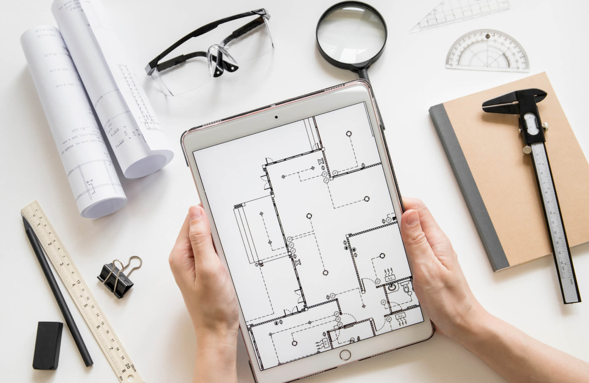 An architect reviewing a digital floor plan on a tablet, surrounded by blueprints, drafting tools, and measuring instruments for a bathroom renovation.