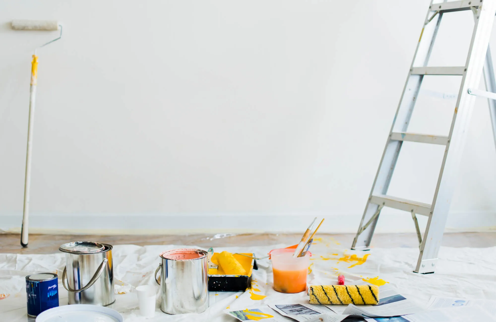 A diy bathroom remodel scene in progress shows a freshly painted white wall with a ladder nearby, surrounded by paint cans, rollers, brushes, and splashes of yellow paint laid out on protective drop cloths.