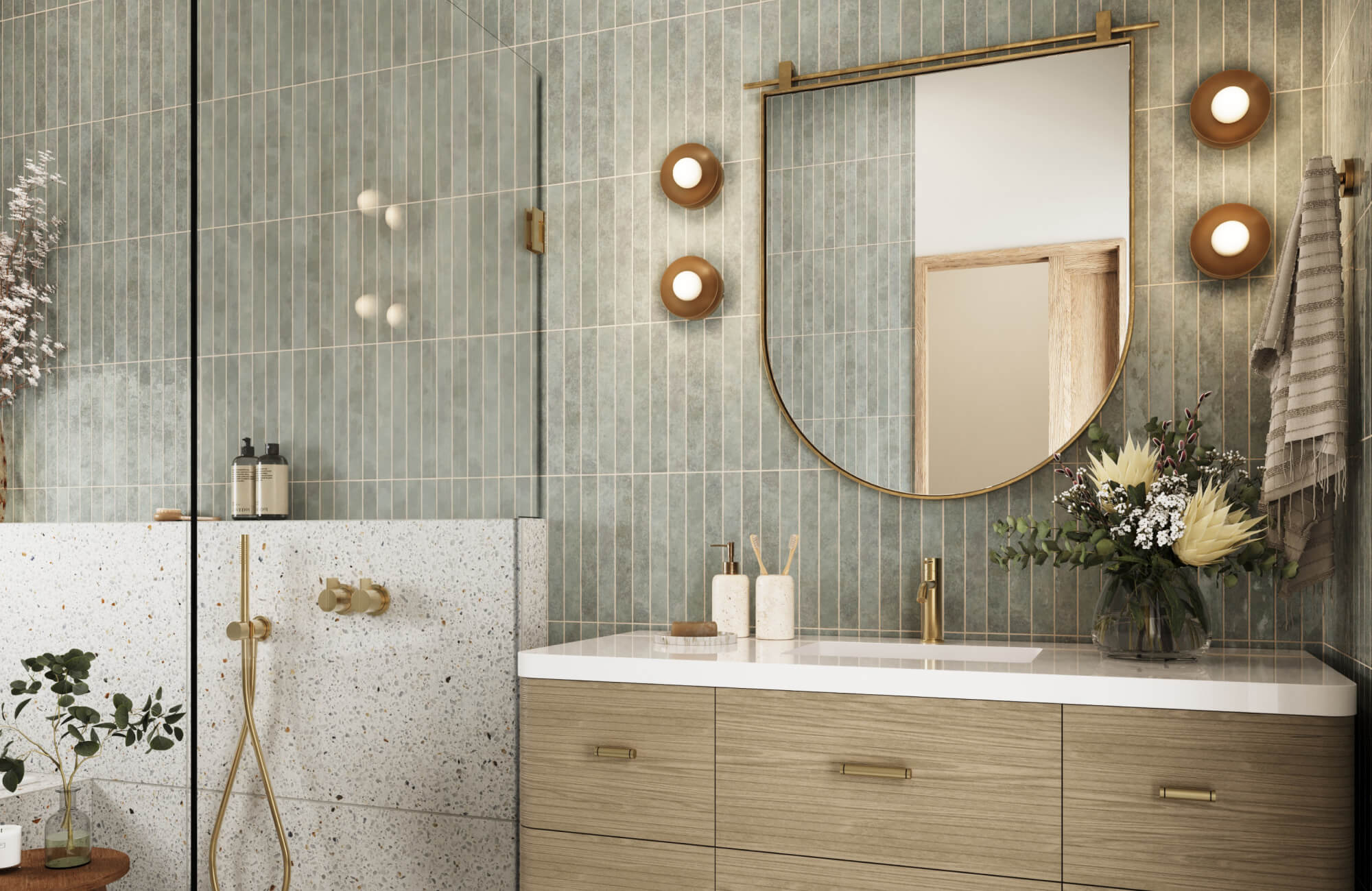 A beautifully styledbathroom featuring soft green vertical tile walls, a wood-front vanity with a crisp white countertop, brass fixtures, warm wall sconces, and a glass-enclosed shower for a calm, modern bathroom retreat.