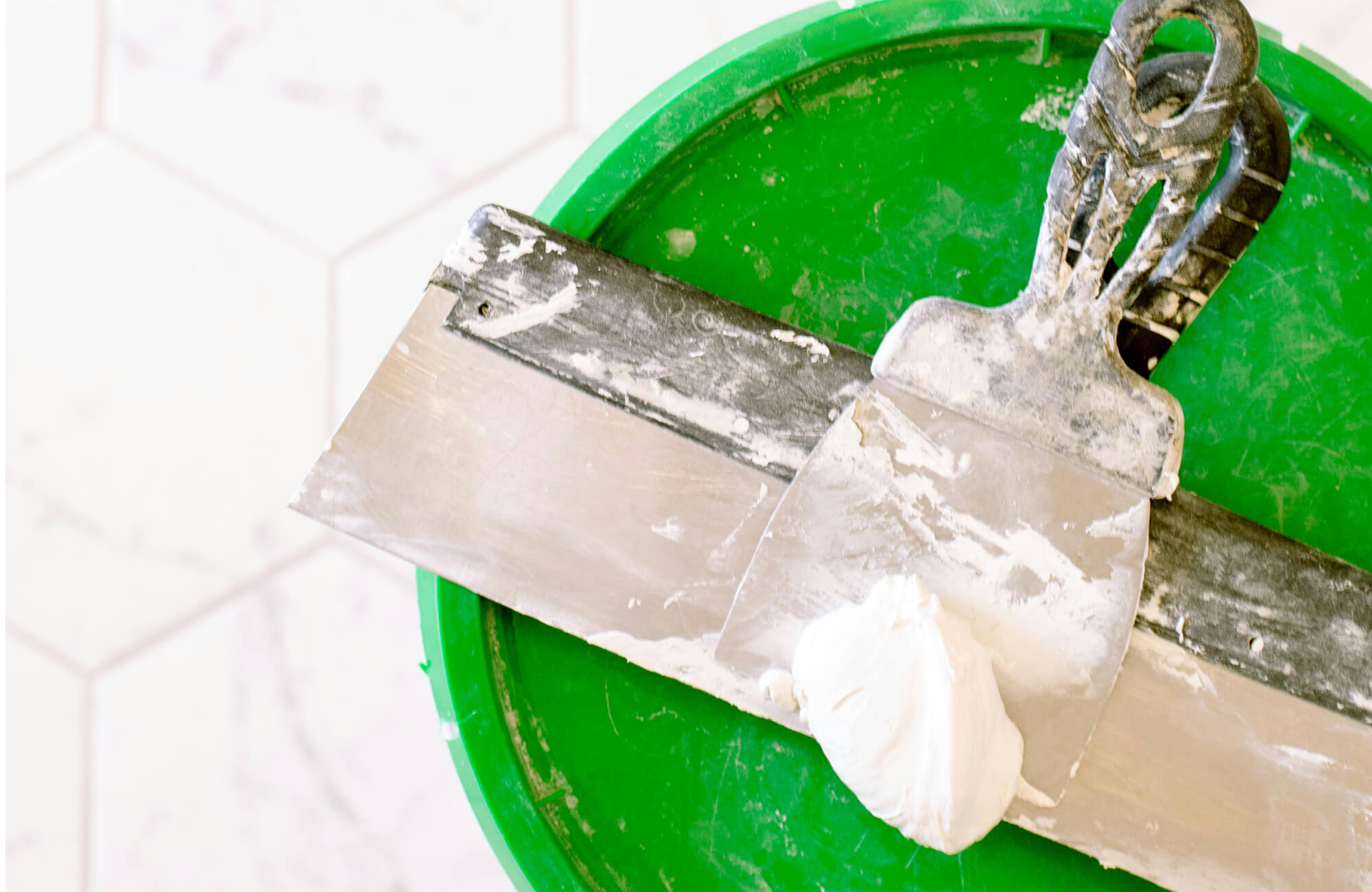 A diy bathroom remodel detail shows putty knives coated with joint compound resting on a green bucket, capturing the messy, hands-on prep work involved in updating and repairing bathroom surfaces.