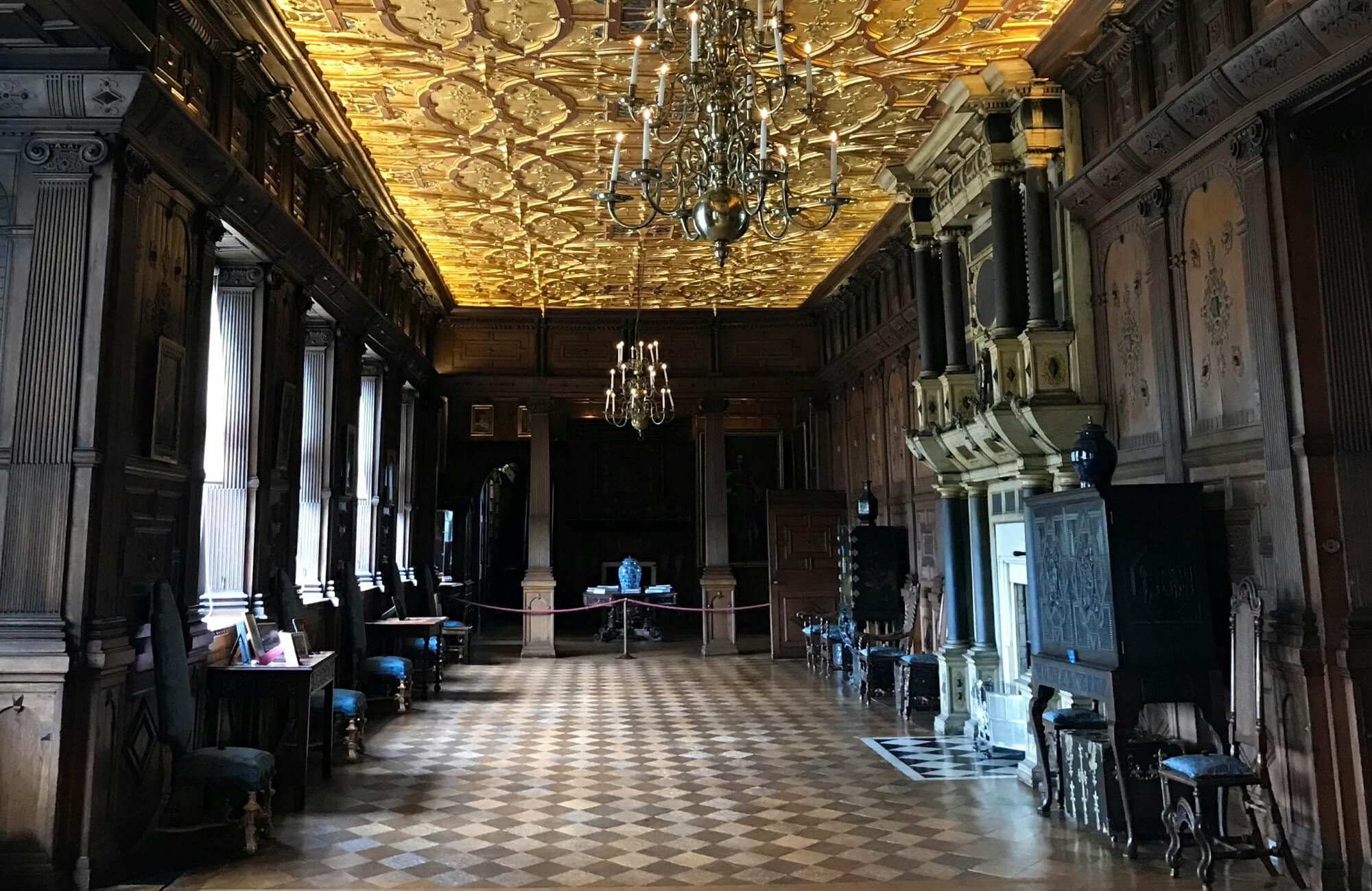 Opulent interior with intricate golden ceiling and vintage checkerboard flooring, featuring dark wood paneling and antique furnishings.