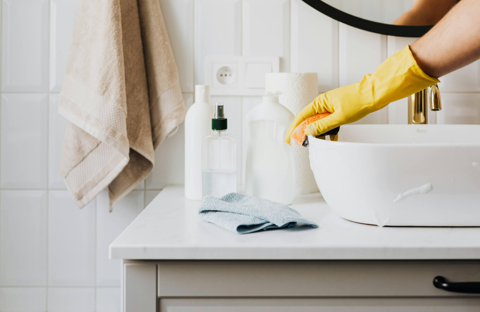 null Close-up of bathroom sink cleaning, with cleaning supplies, folded towel, and beige hanging towels against a tiled backsplash.