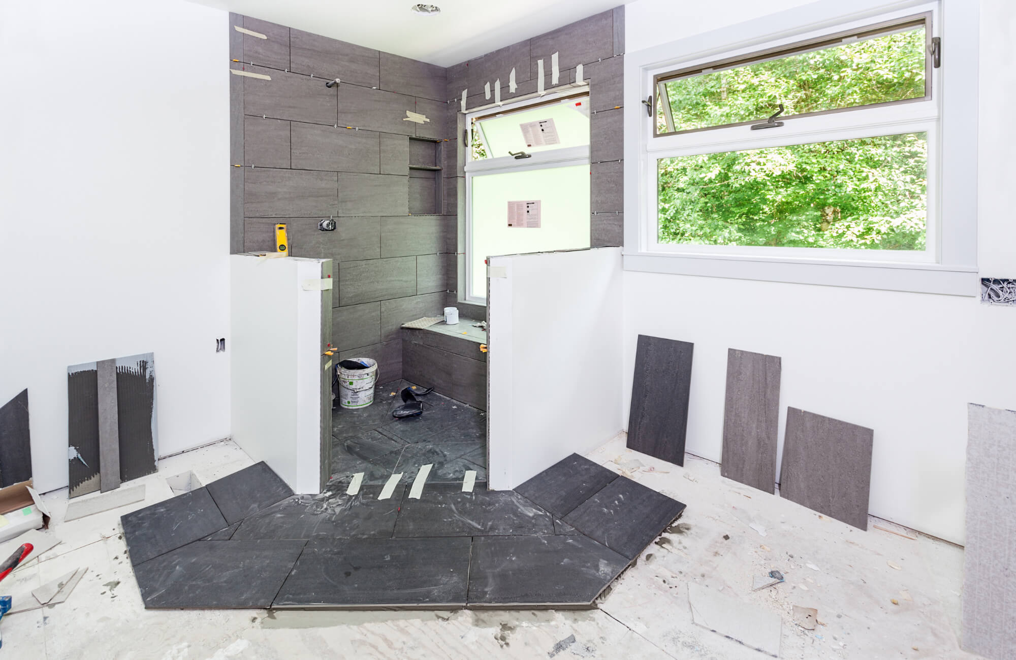 Bathroom under construction featuring large-format gray ceramic tiles being installed in a walk-in shower with a bench and window view.