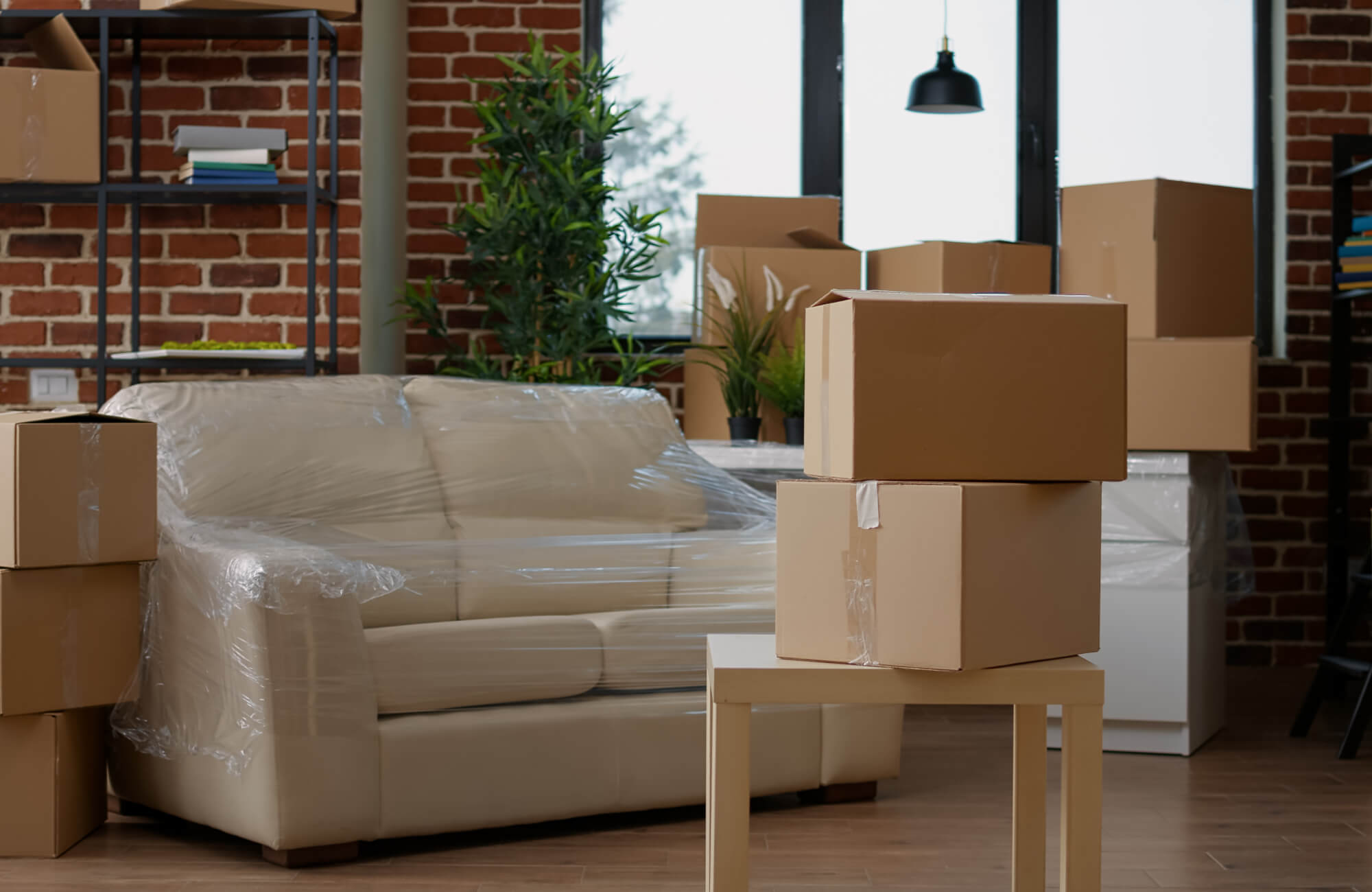 null Inside a loft living room, brown moving boxes sit on a light wood floor beside a cream sofa wrapped in plastic, with red brick walls and green plants in the background.