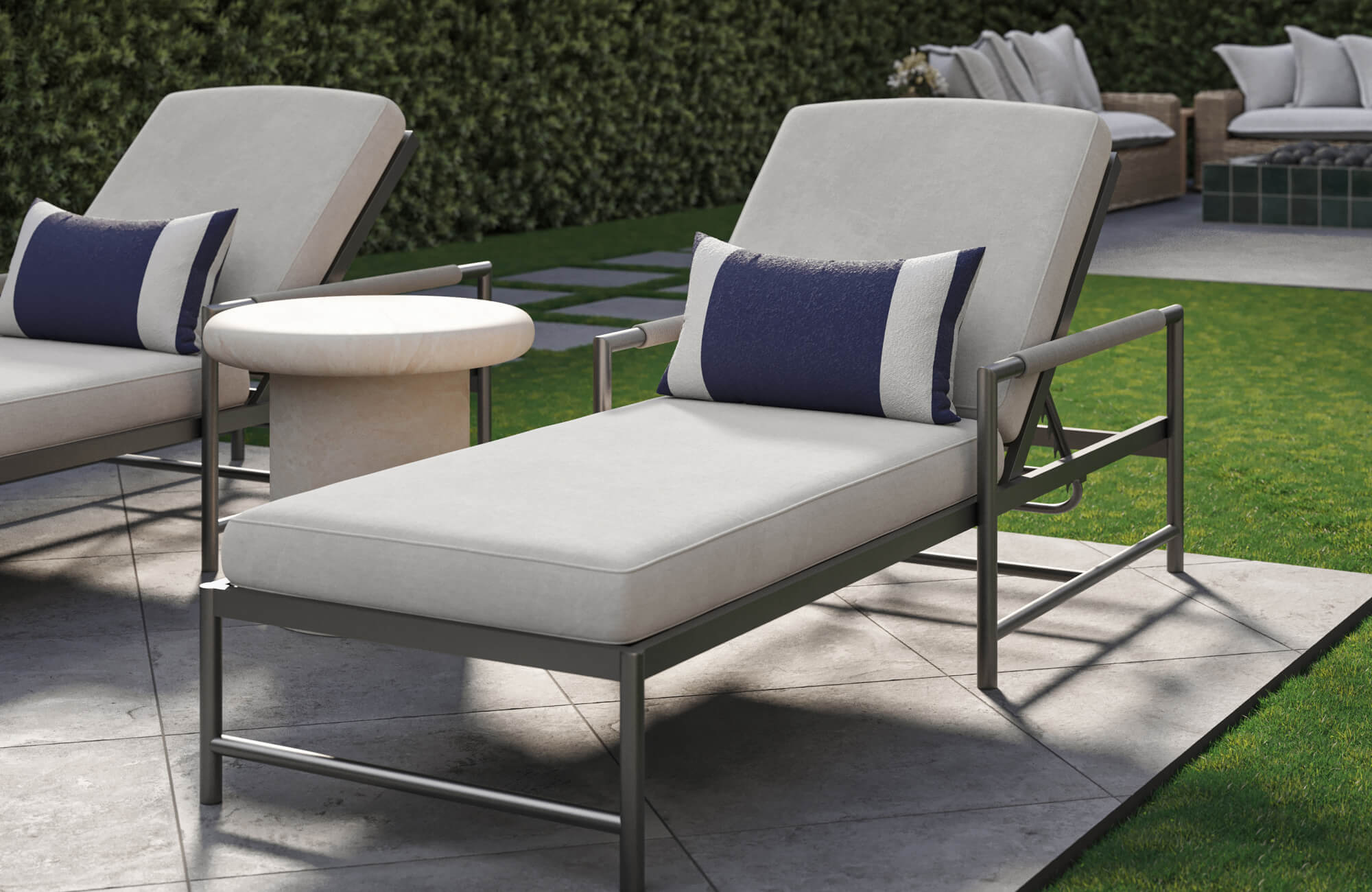 null Beside a green lawn, a gray metal lounge chair with light gray cushions and a navy blue pillow sits on a light stone patio next to a small beige side table.