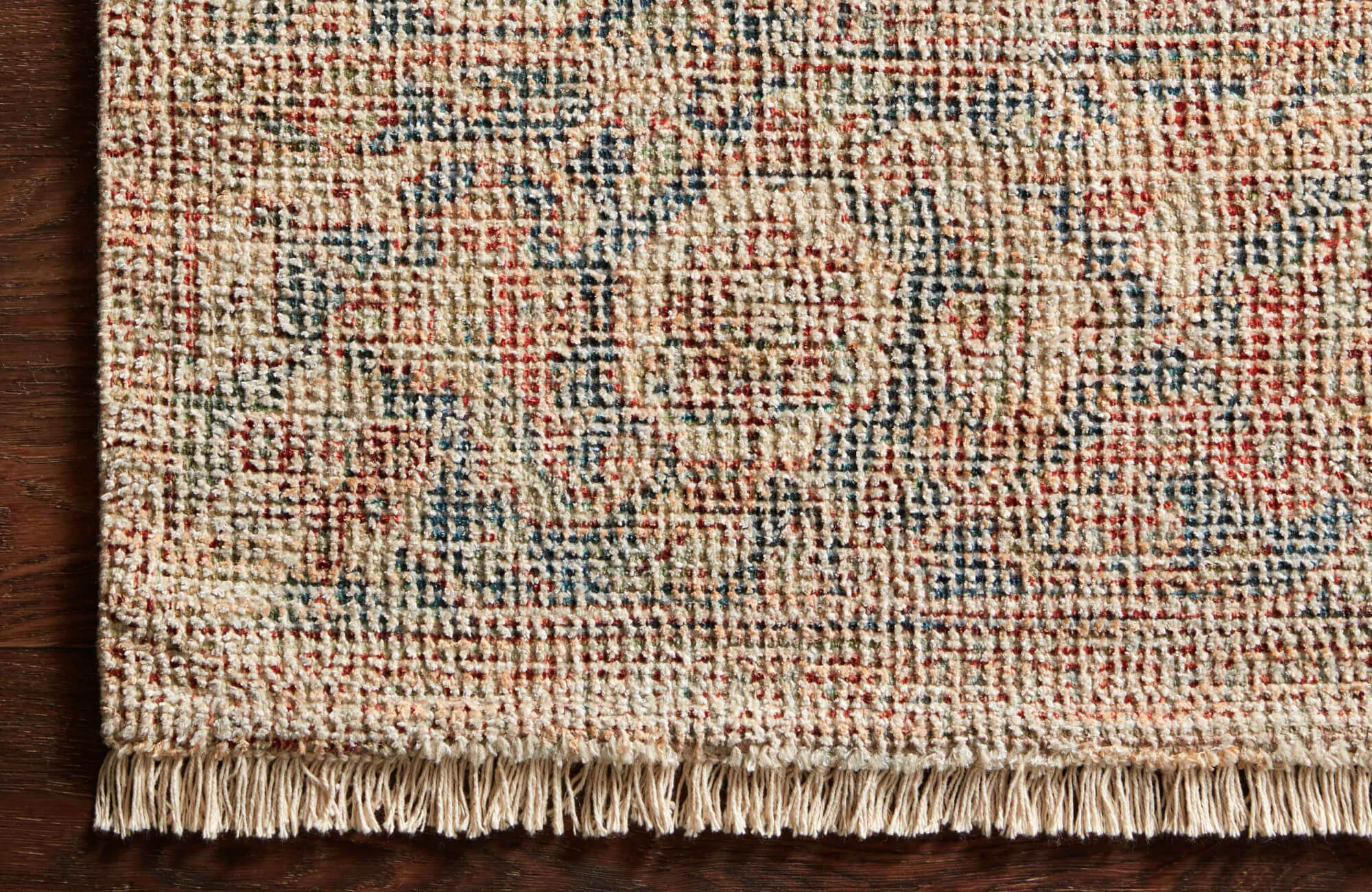 Textured rug corner featuring a multicolor floral pattern and knotted fringe border, styled over hardwood flooring.