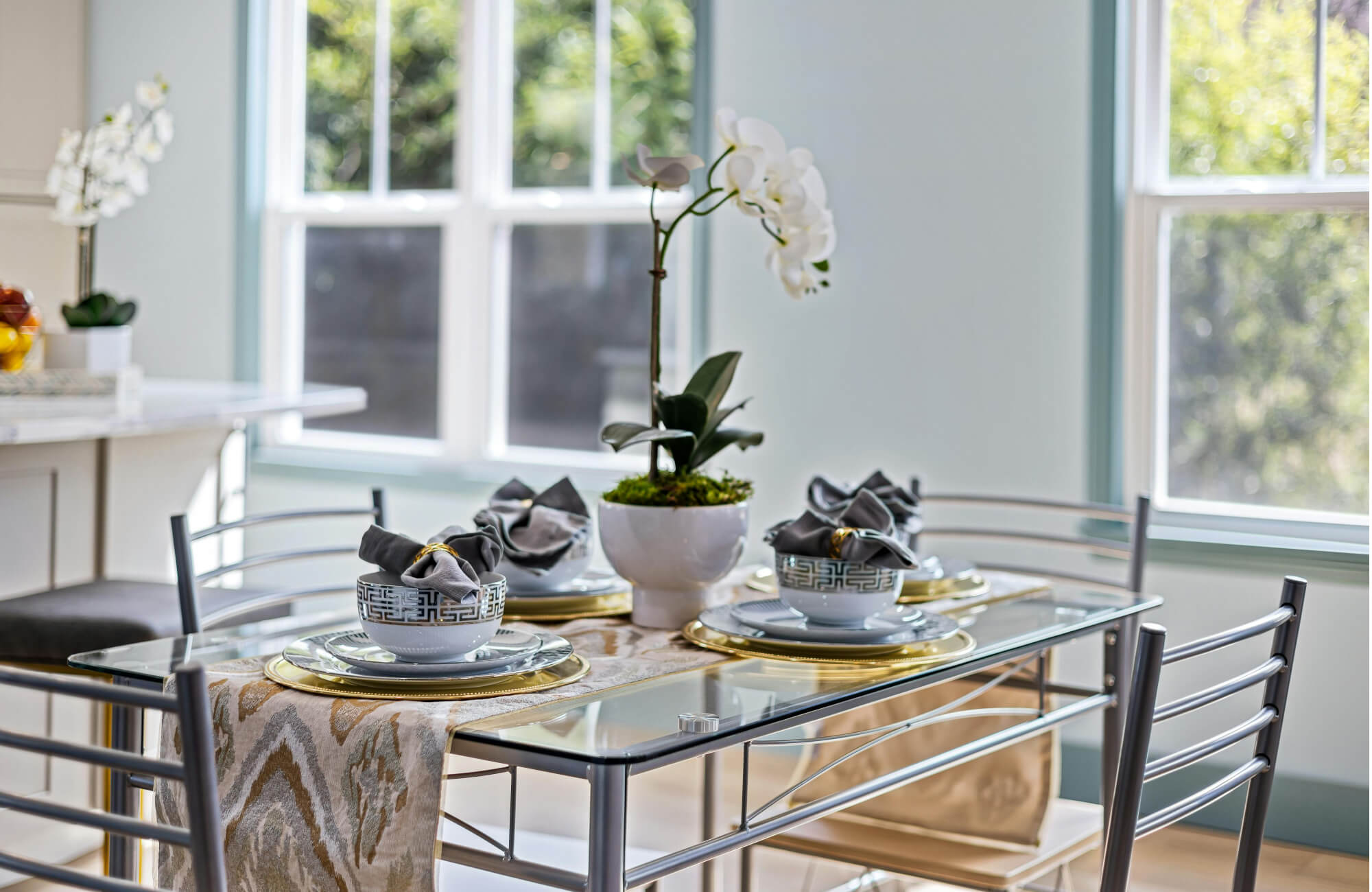 null A bright, elegant dining nook styled around a sleek glass dining table, accented with gold-rimmed place settings, soft gray linens, and a sculptural orchid centerpiece that enhances the space’s light-filled, modern charm.