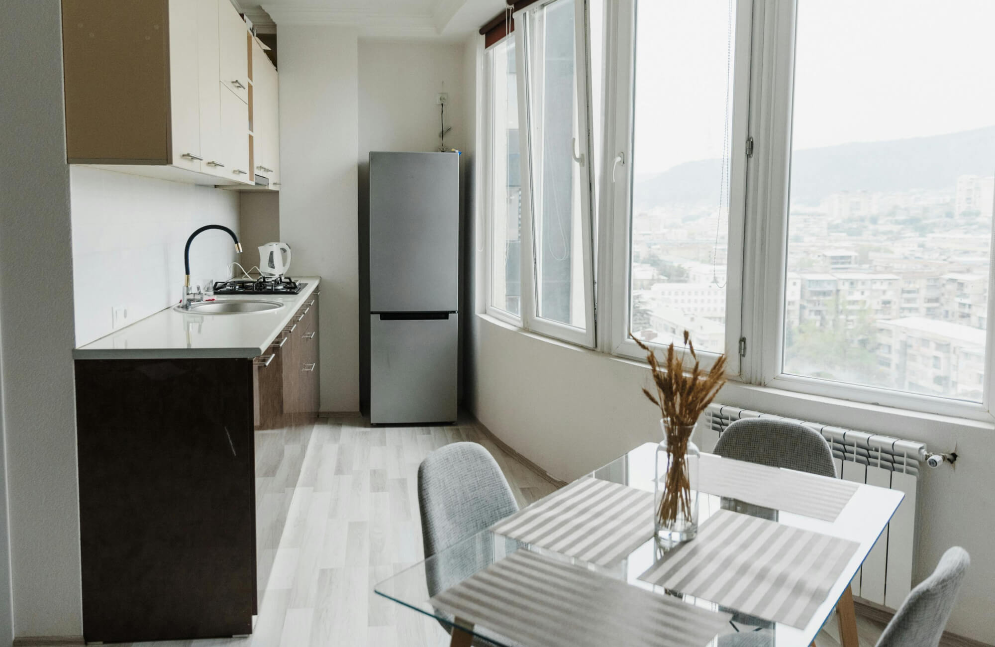 null A compact, light-filled kitchen-dining space features a minimalist glass dining table that visually opens the room, paired with soft gray chairs, clean-lined cabinetry, and expansive windows that enhance the modern, airy feel.