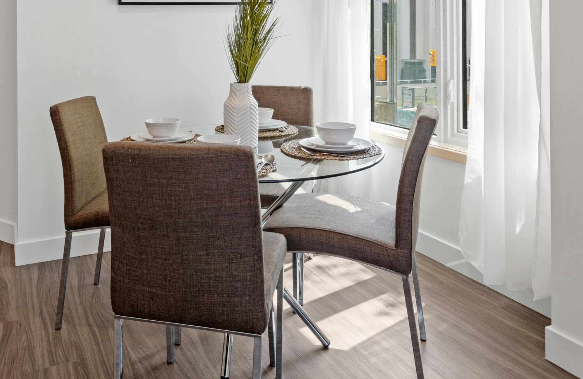 null A cozy, light-filled dining corner highlights a round glass dining table paired with upholstered chairs, where soft neutral tones, woven placemats, and a simple vase centerpiece create a warm, contemporary feel.