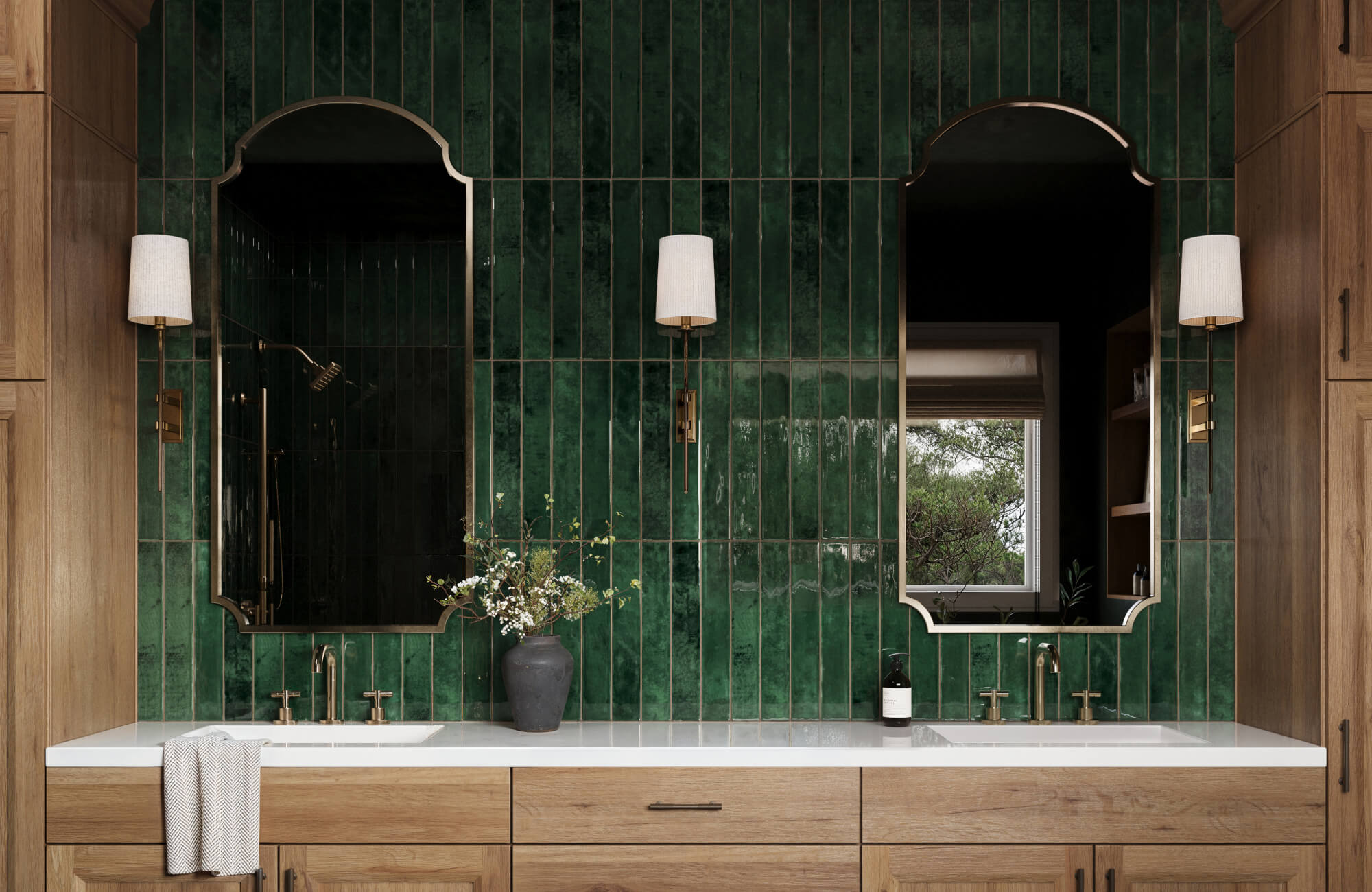 Rich green vertical tile backsplash highlights a double sink vanity with brass fixtures, natural wood cabinets, and sculpted mirrors in a modern bath.