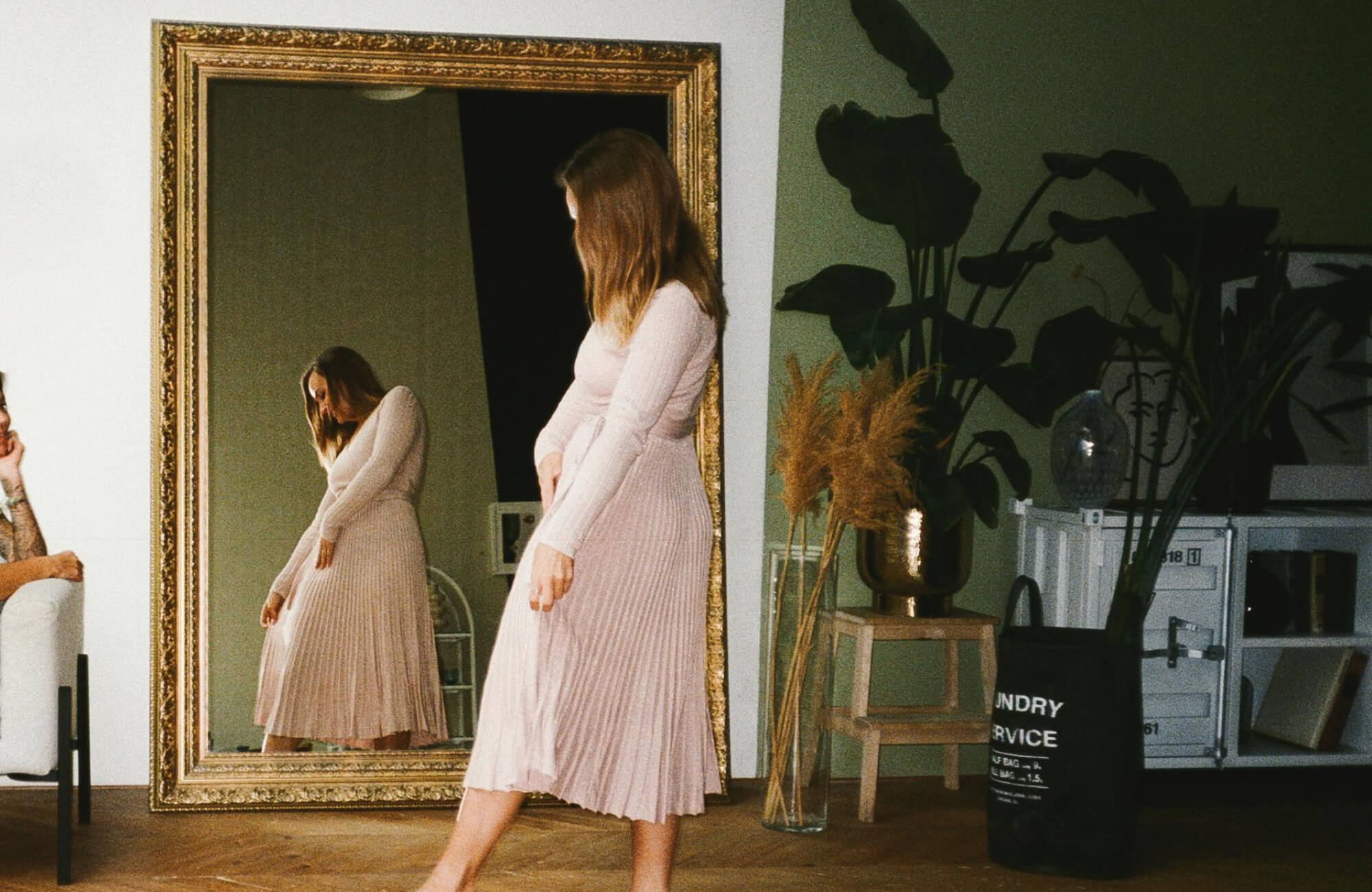 null A spacious, softly lit room featuring a large floor mirror with an ornate gold frame, reflecting a woman in a pleated dress and surrounded by lush indoor plants and warm natural décor accents.