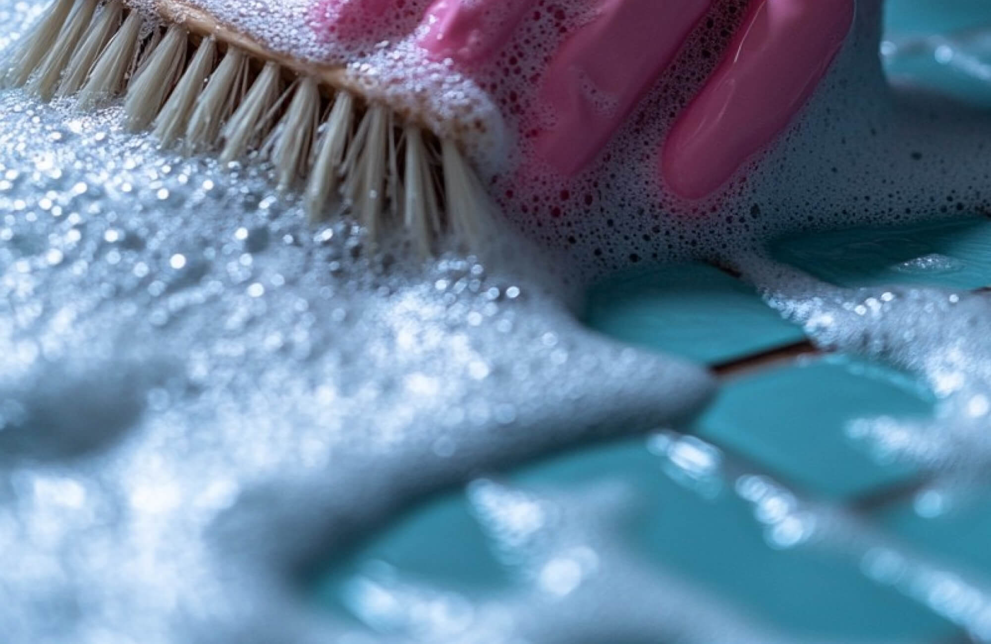 A pink-gloved hand scrubbing mosaic tiles with a bristle brush, creating a frothy layer of soap bubbles for a deep clean.