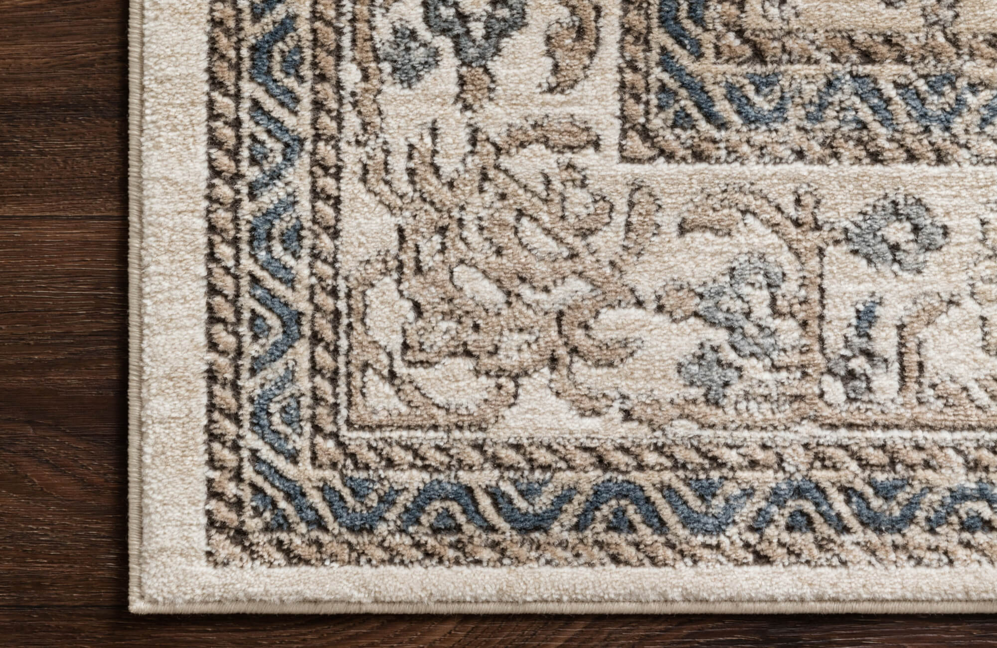Detailed corner of a polypropylene rug with a classic floral and geometric border in beige, brown, and blue tones on dark wood flooring.