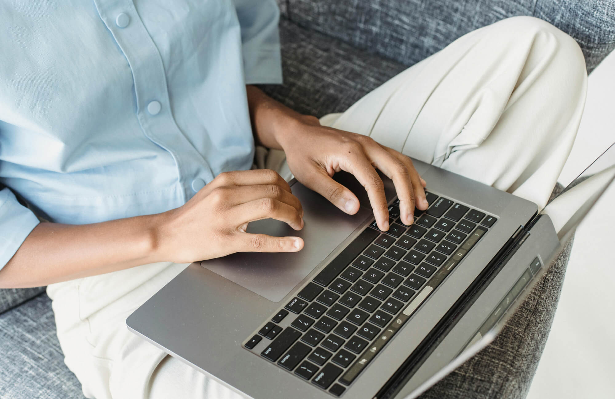 Person in casual attire typing on a laptop while seated on a gray upholstered sofa, suggesting online research or shopping.
