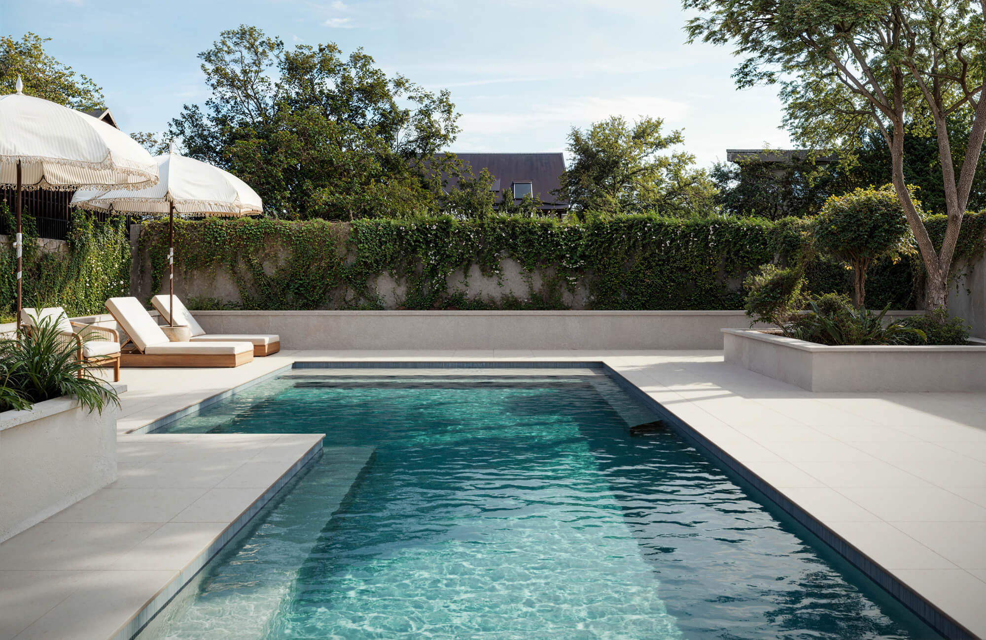 Modern poolscape with smooth ivory porcelain paver tiles, lush greenery, and elegant loungers under fringed umbrellas for serene relaxation