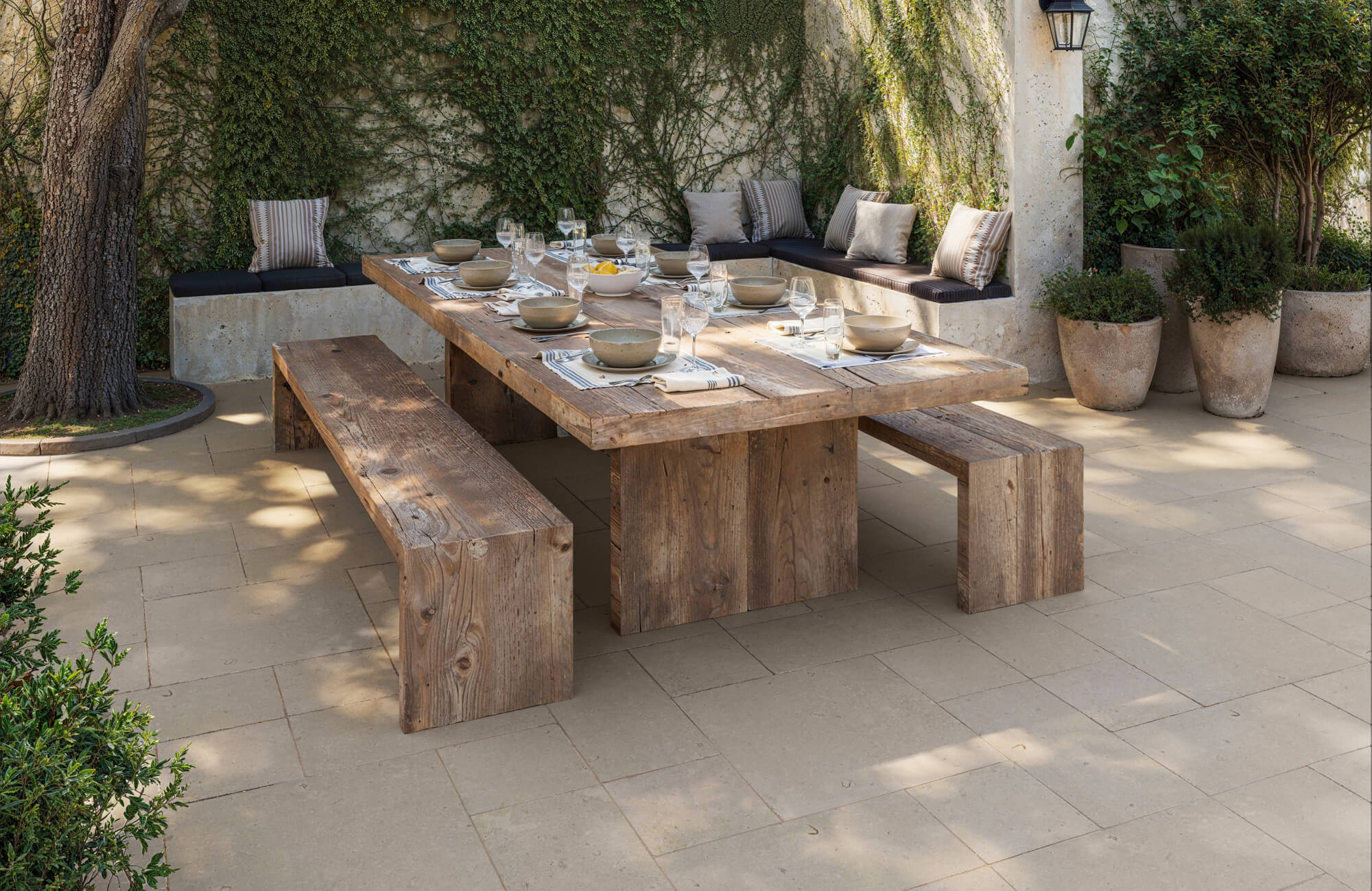 Inviting patio with rustic wood dining set, beige stone look pavers, and built-in bench seating against a vine-covered wall.