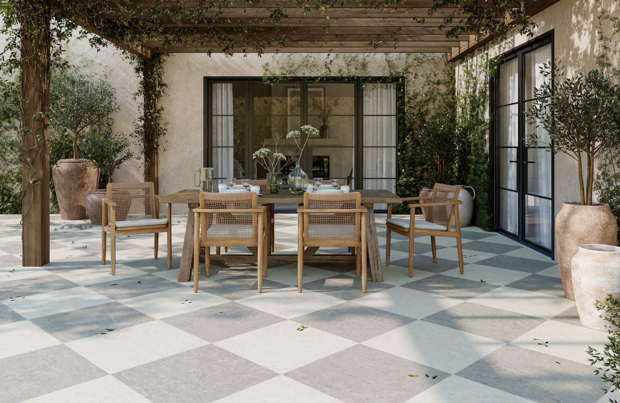 Chic outdoor dining area with checkerboard stone look paver tiles, wooden pergola draped in vines, and natural wood furniture with cane backs.