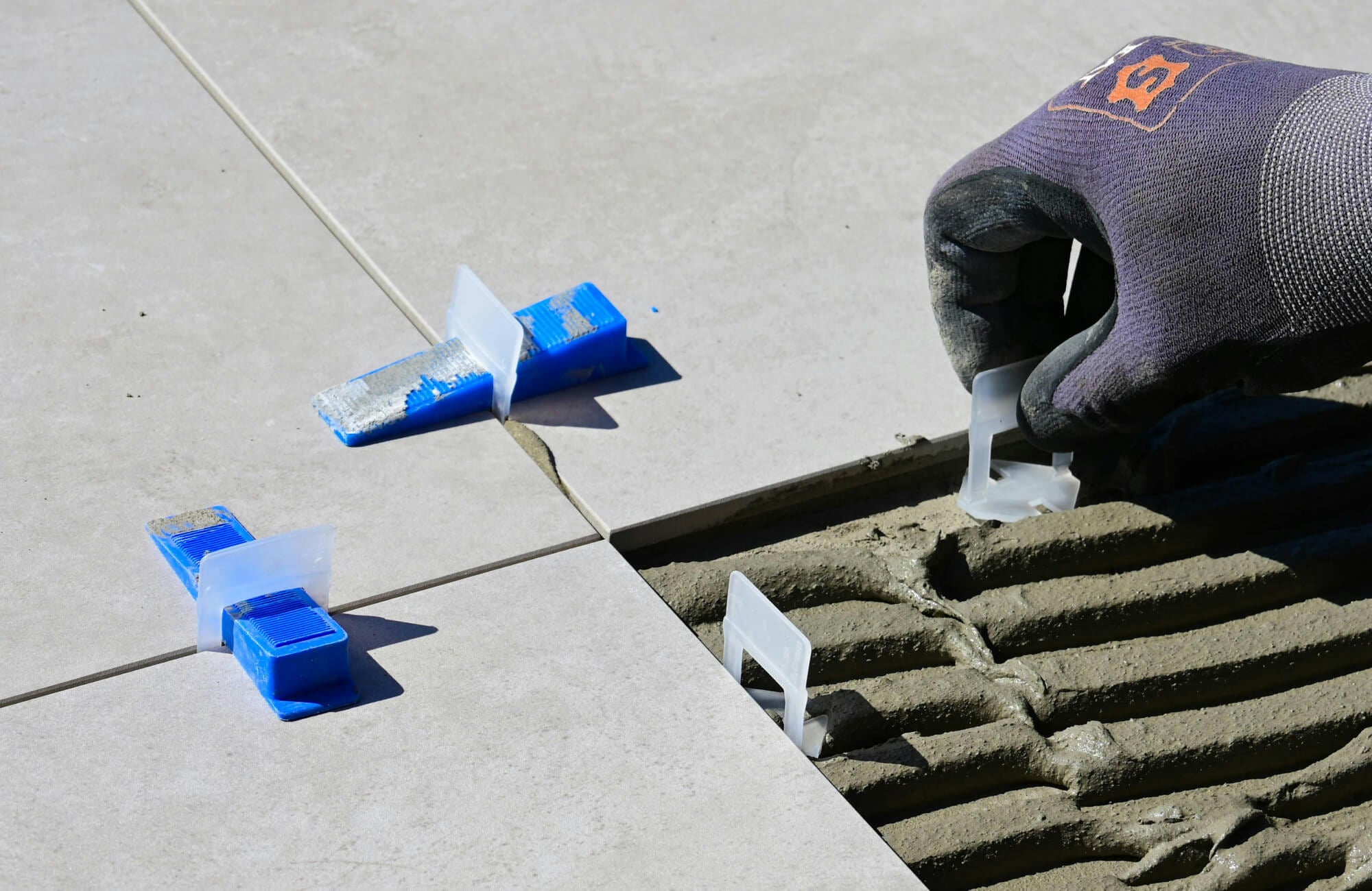 Close-up of paver tile installation using leveling spacers, with a gloved hand setting clips over fresh mortar for precise alignment.