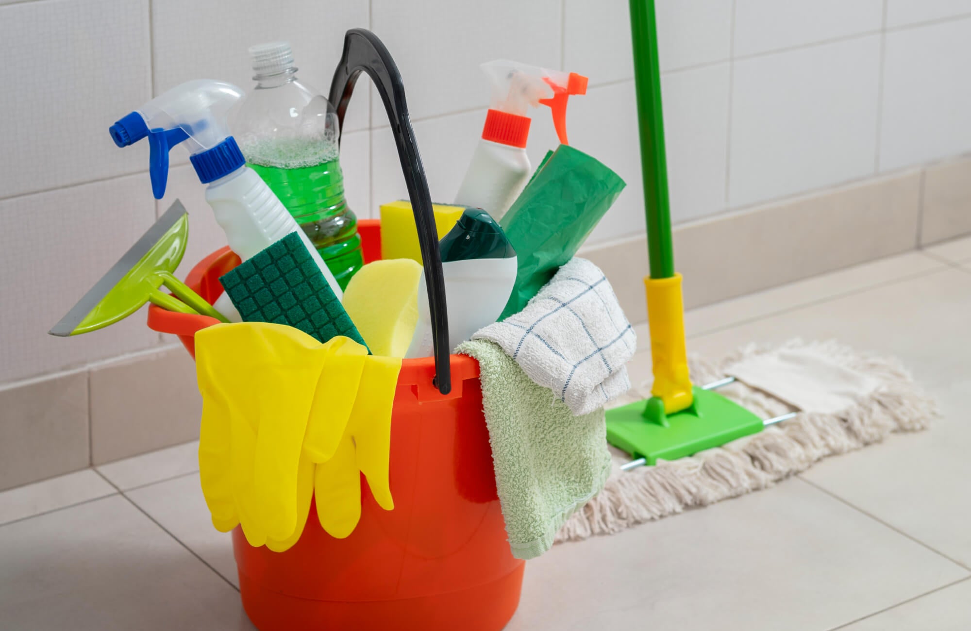 Red bucket filled with outdoor paver cleaning supplies including spray bottles, gloves, sponges, scrub pads, and a mop on tiled surface.