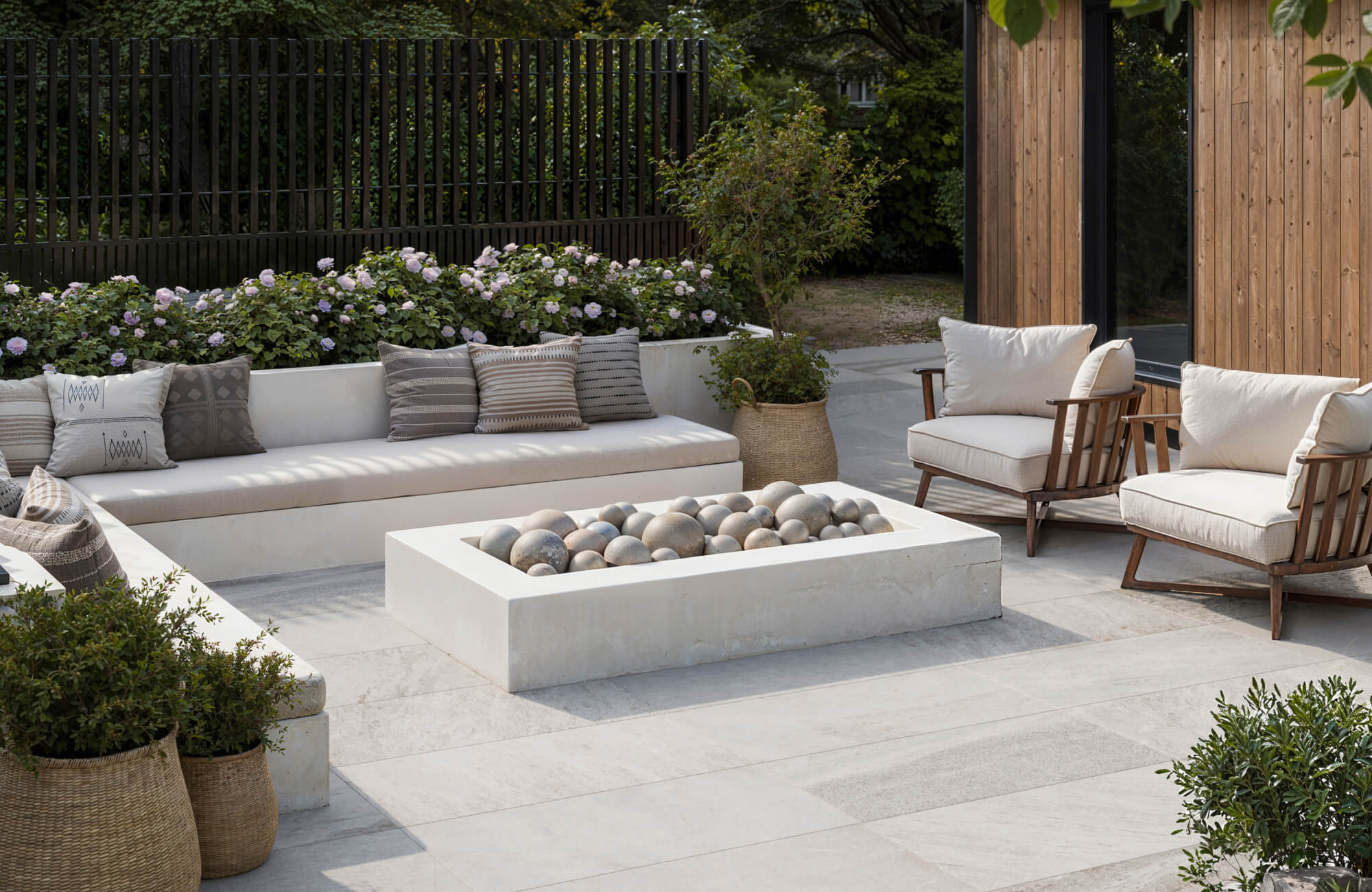 Modern patio with light stone look pavers, built-in seating with neutral cushions, a sculptural fire pit, and cozy wooden lounge chairs.