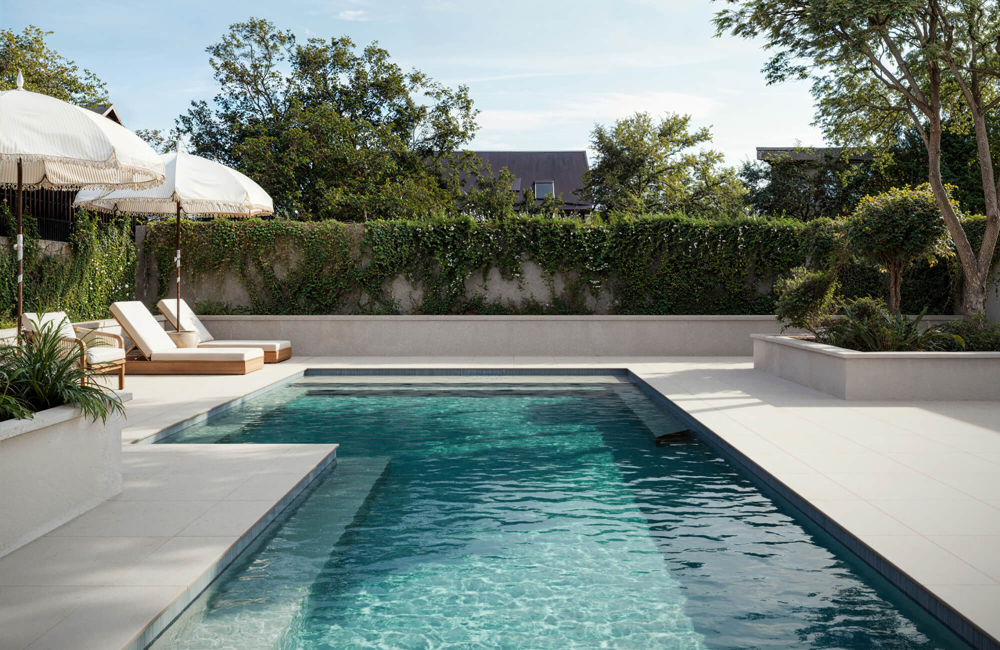Elegant poolside with light porcelain pavers, crisp lounge chairs under white umbrellas, and lush greenery along a vine-covered wall.