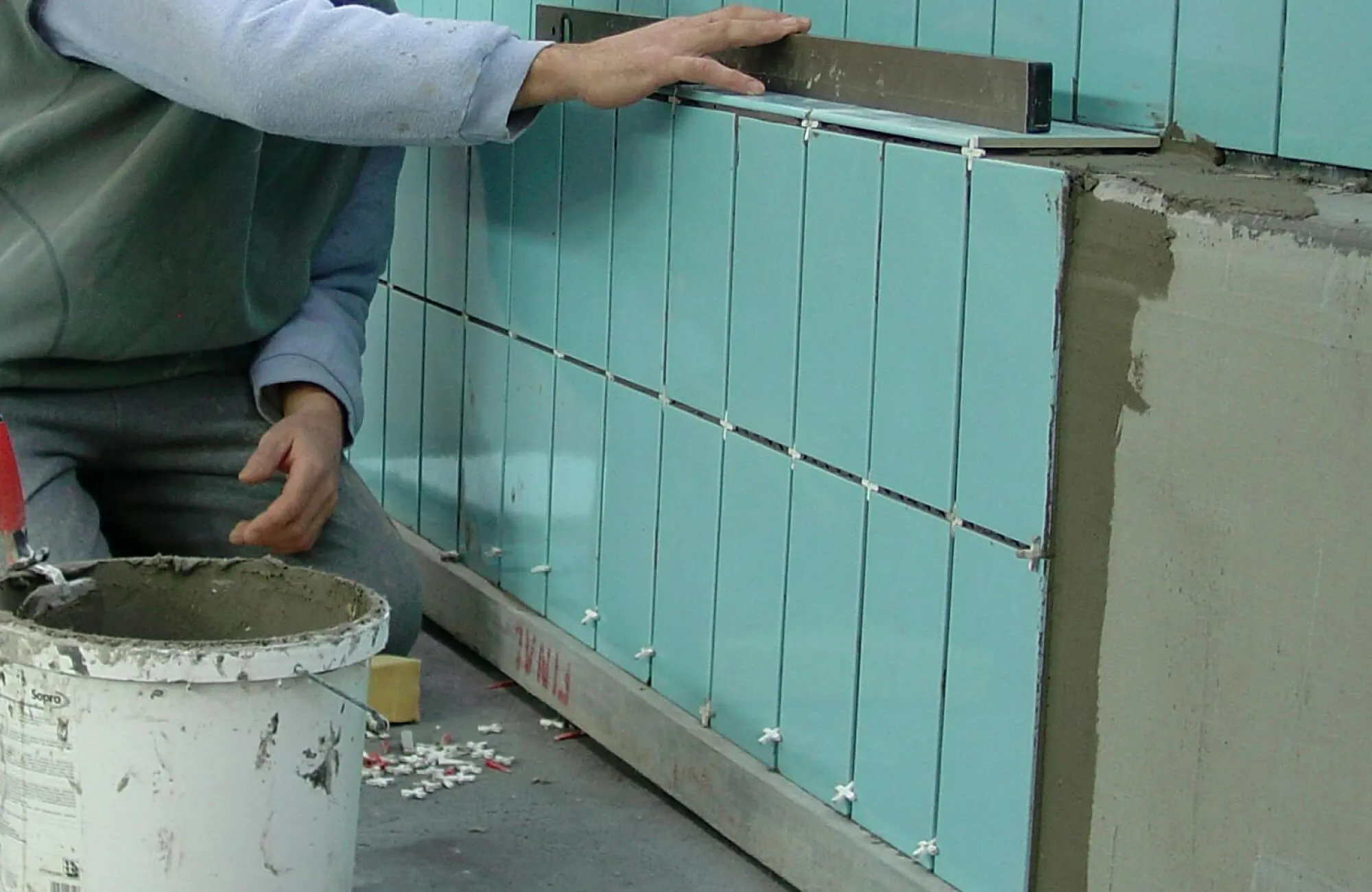 Skilled worker aligning glossy aqua-blue subway tiles on a wall, with precision tools and an adhesive bucket nearby, creating a sleek modern finish.