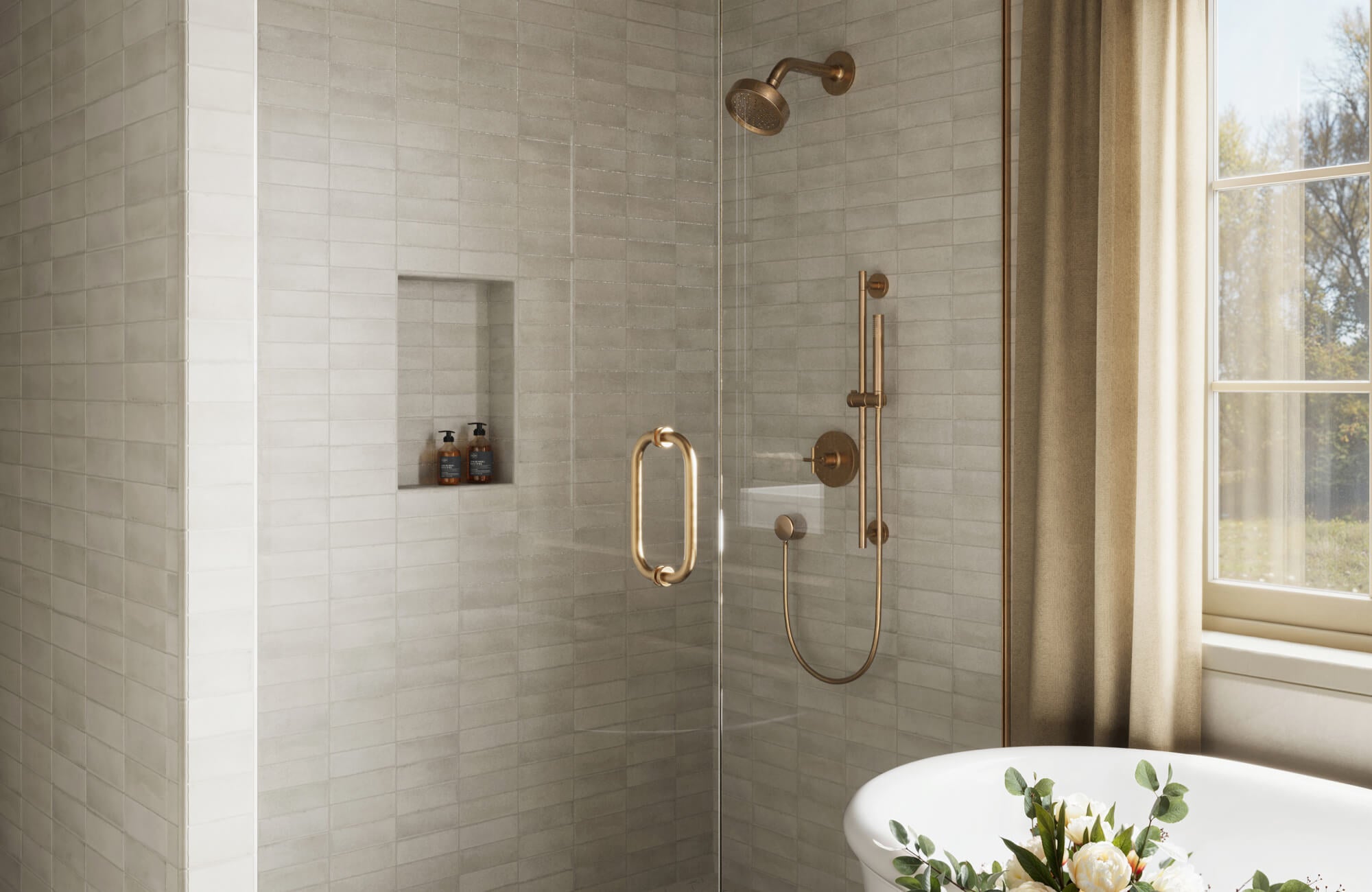 Soft gray subway tiles line a walk-in shower with brushed brass fixtures and a built-in niche beside a sunlit soaking tub
