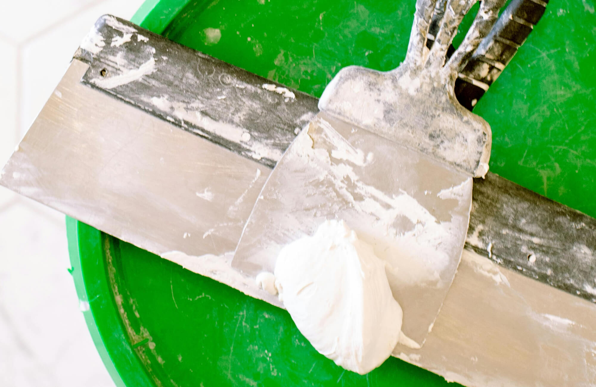 Smooth metal trowel with a dollop of white grout rests on a green bucket, showcasing tools for tile installation.