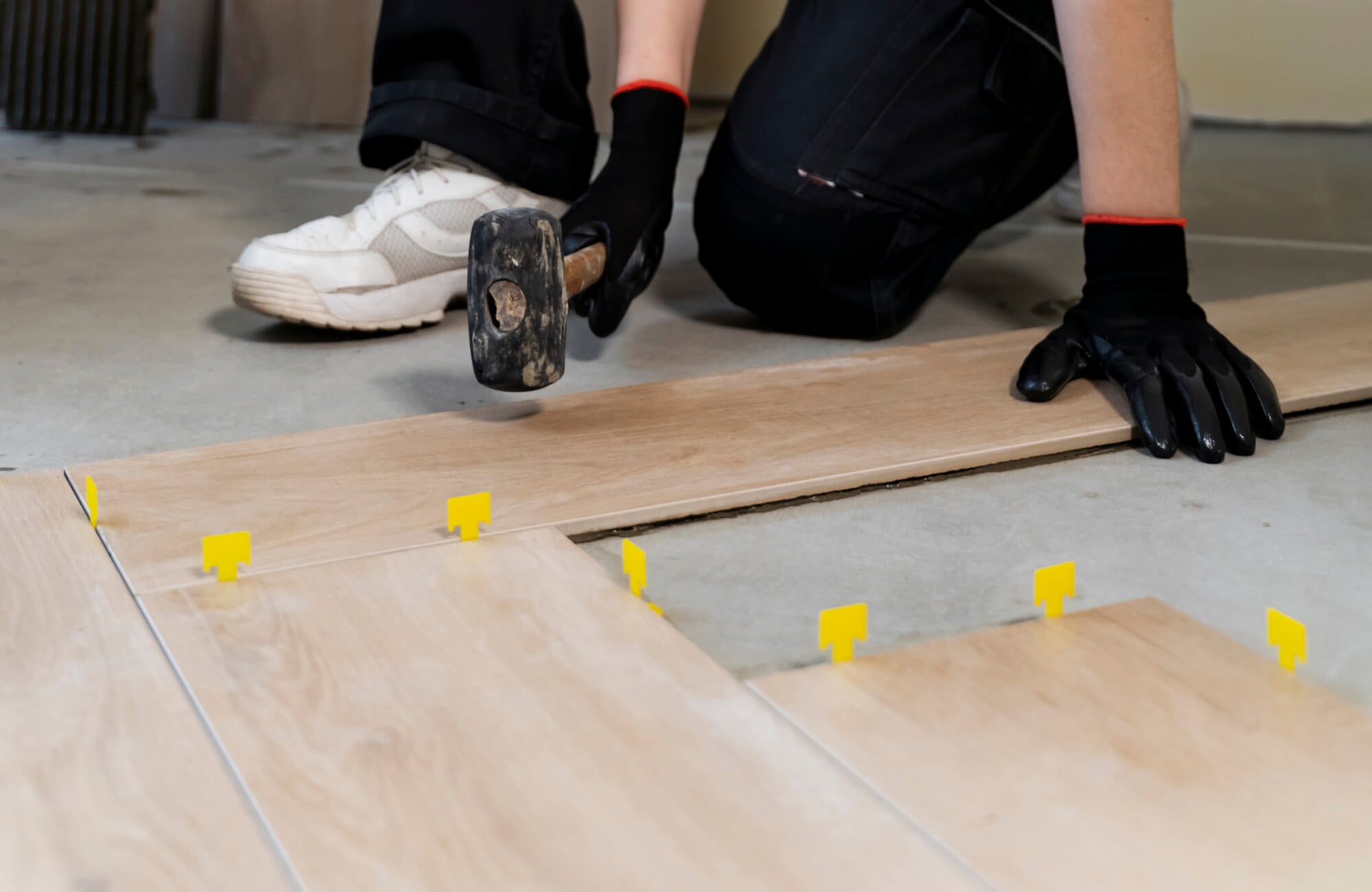 Worker aligns wood look tiles with yellow spacers, using a rubber mallet for precise installation on a smooth surface.