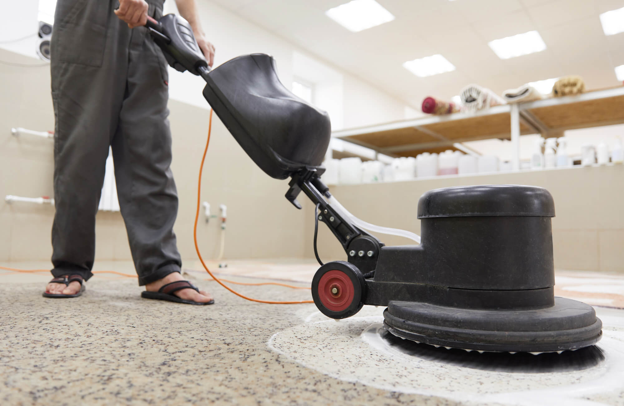 Worker operating a commercial floor cleaning machine on carpet in a professional rug cleaning facility with rugs and supplies in the background.