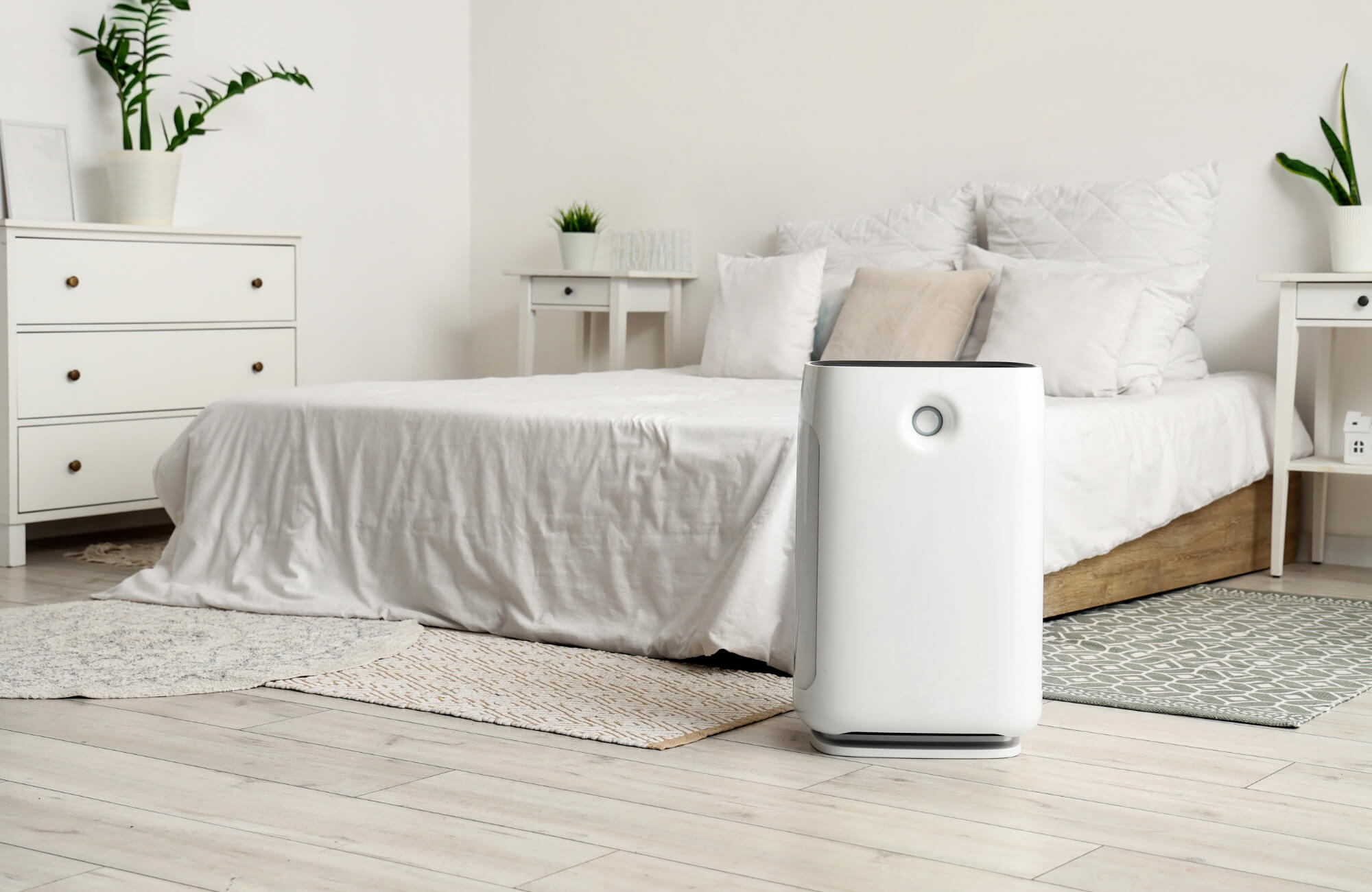 Modern bedroom with white bedding, wood furniture, potted plants, small rugs on a light floor, and a sleek air purifier in the foreground.