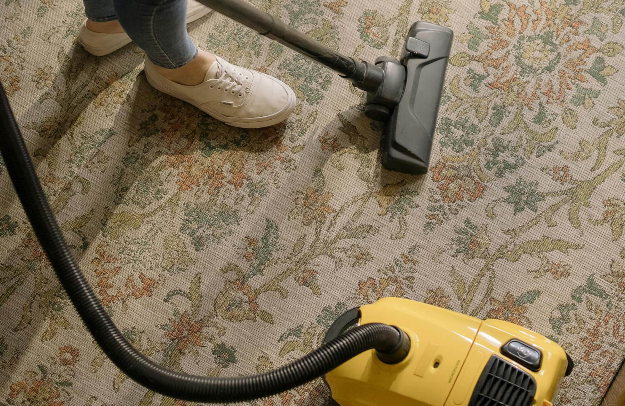 Person vacuuming a patterned wool rug with a yellow canister vacuum, wearing casual jeans and white sneakers.