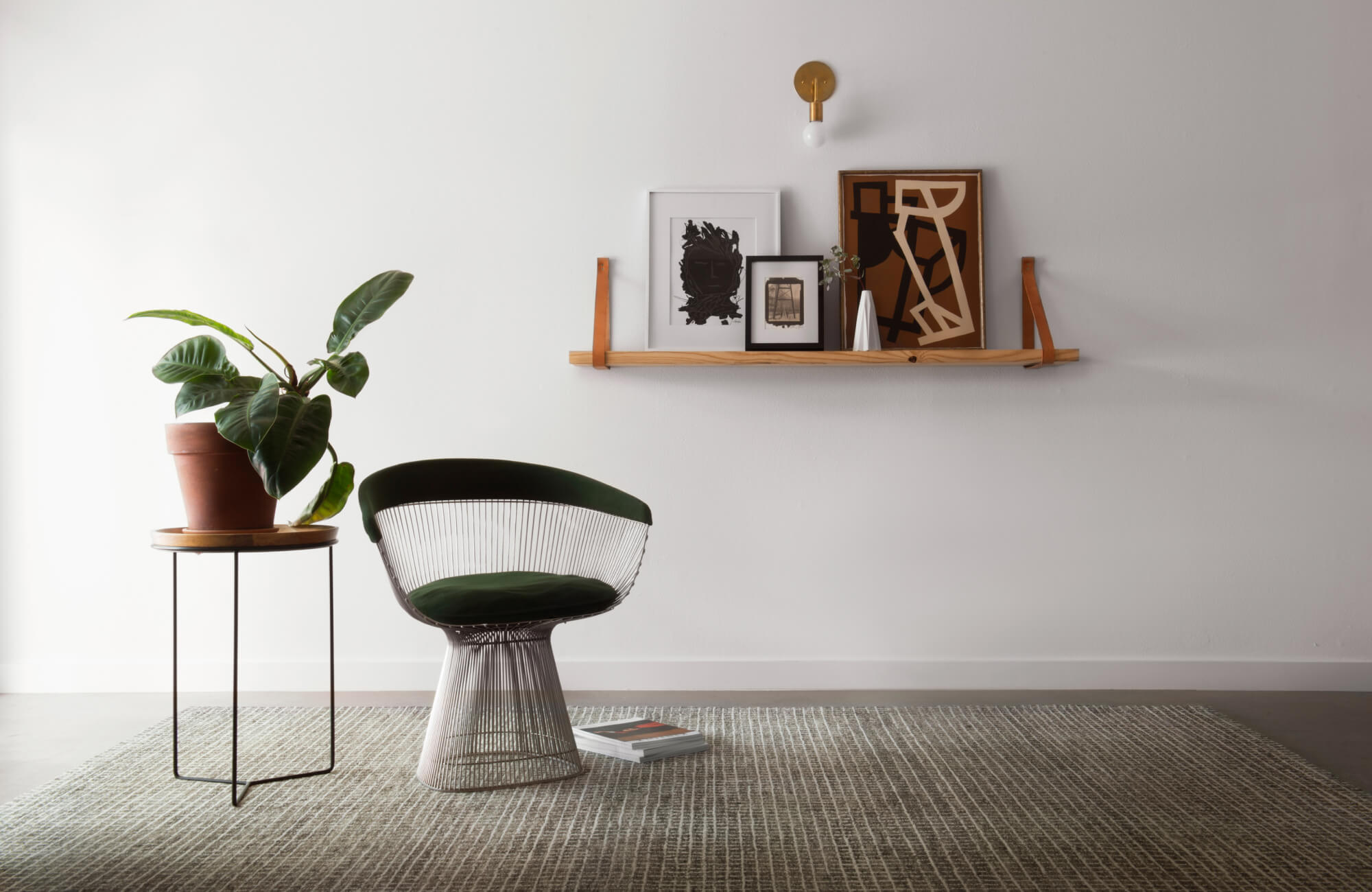 Minimalist room with a wool rug, a small side table holding a potted plant, and a floating shelf displaying framed art and decor against a clean white wall.