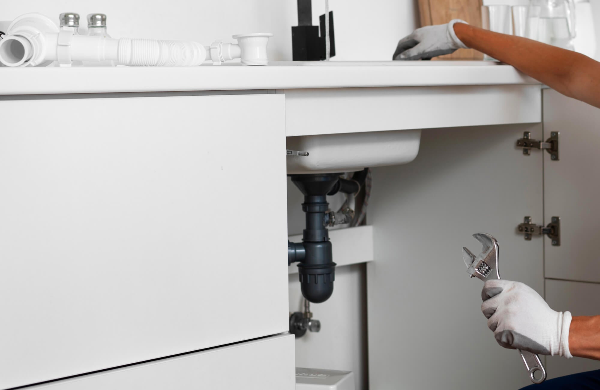 null Close-up of a plumber repairing a bathroom double vanity sink, showing gloved hands holding a wrench beneath the countertop with visible piping and plumbing tools arranged nearby.