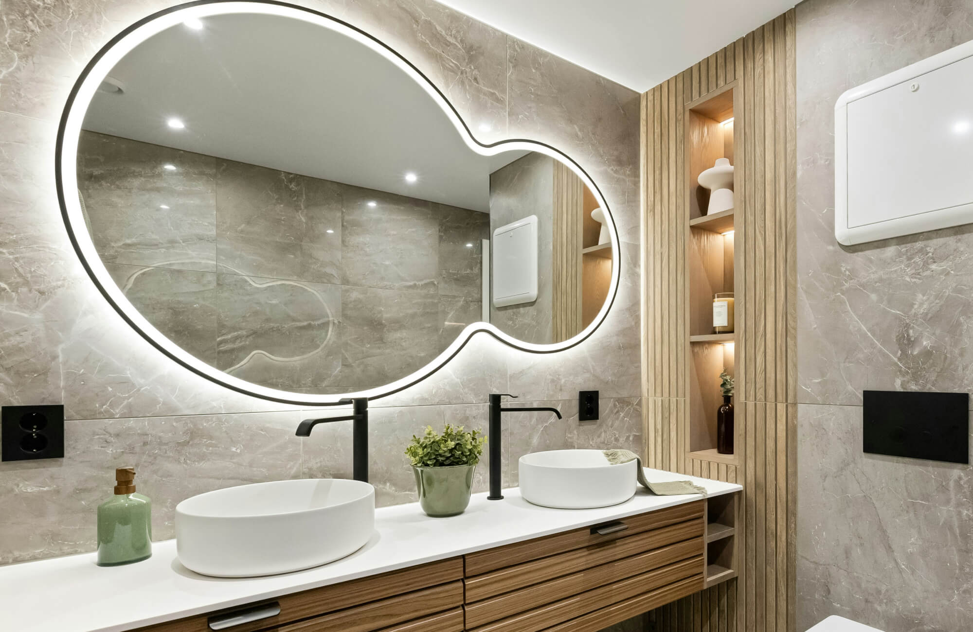 null Contemporary bathroom double vanity featuring dual vessel sinks, matte black faucets, a sculptural backlit mirror, and warm wood cabinetry accented by marble walls for a sleek and sophisticated aesthetic.
