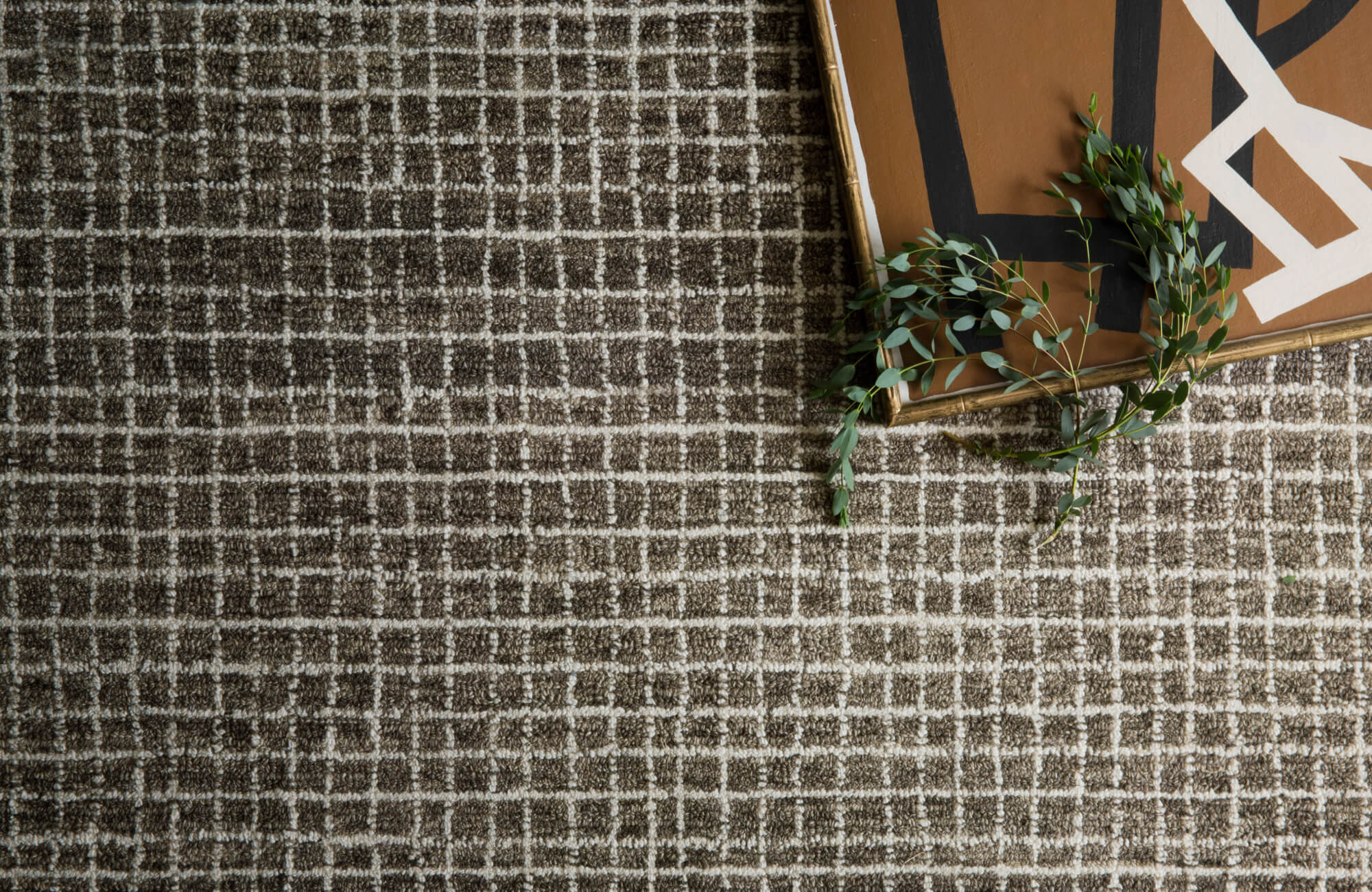 Close-up of a textured brown rug with a subtle grid pattern, accented by a decorative tray and greenery for a modern, earthy look.