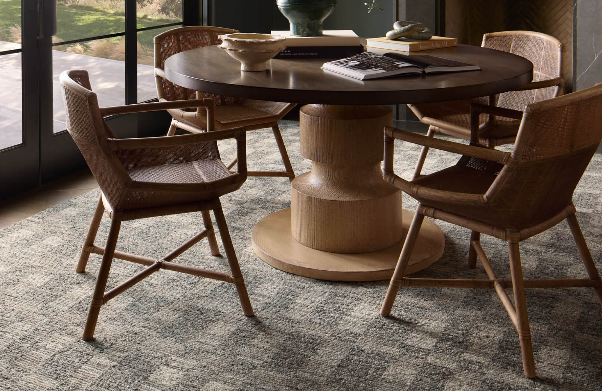 Warm dining space with woven chairs and a round wooden table set atop a patterned brown rug that adds depth and organic texture.