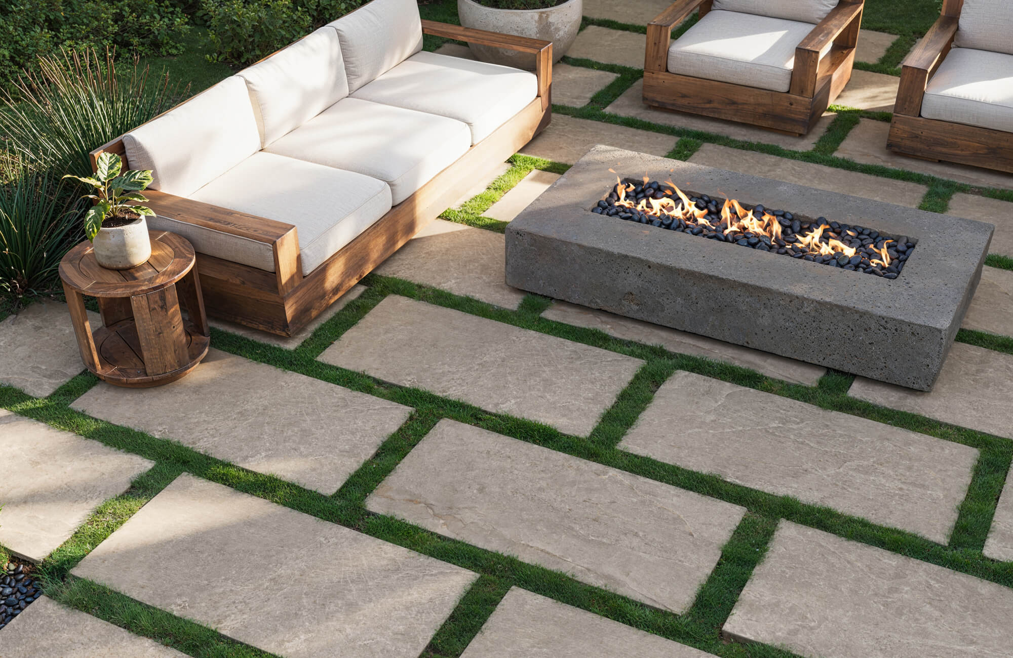 null Outdoor patio with stone look porcelain pavers set in grass, featuring wood furniture and a modern fire pit centerpiece.