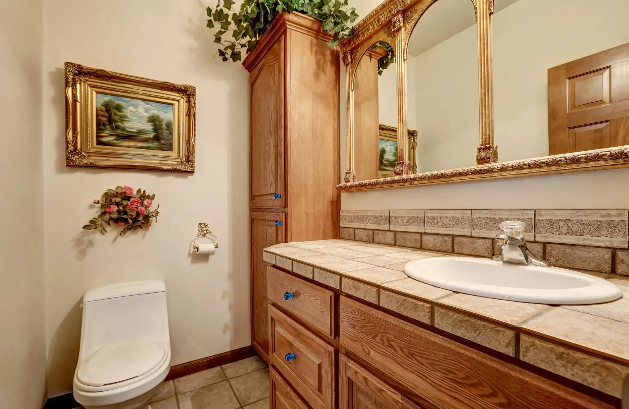 Traditional bathroom with a wood vanity, tiled countertop, ornate framed mirror, wall art, and decorative greenery.