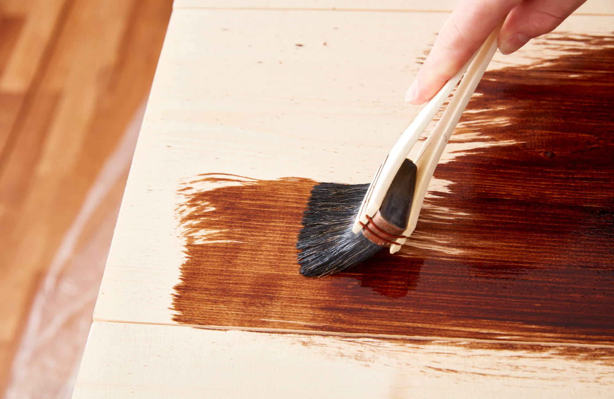 Close-up of a hand applying dark wood stain with a brush on a light wooden surface, highlighting the finishing process.