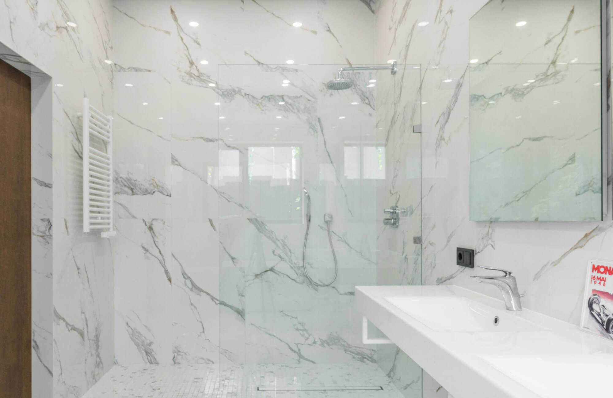 Marble bathroom featuring large-format white marble walls with gray veining, a frameless glass shower enclosure, floating white vanity with integrated sink, and sleek chrome fixtures for a clean, modern aesthetic.