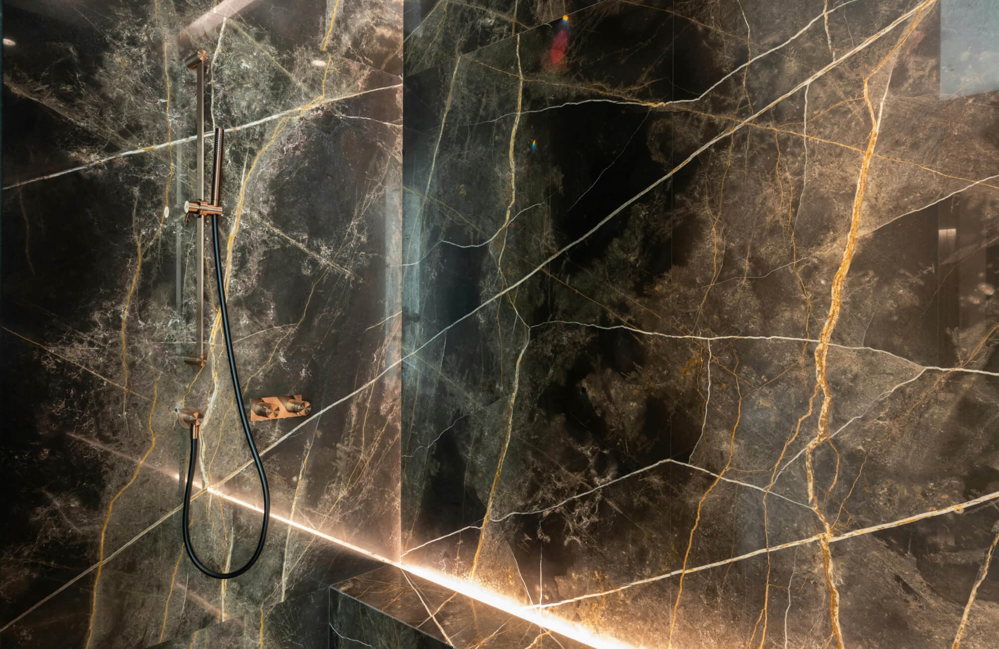 Marble shower clad in dramatic black marble with bold white and gold veining, featuring sleek bronze fixtures and integrated linear lighting for a luxurious, high-contrast modern bathroom design.