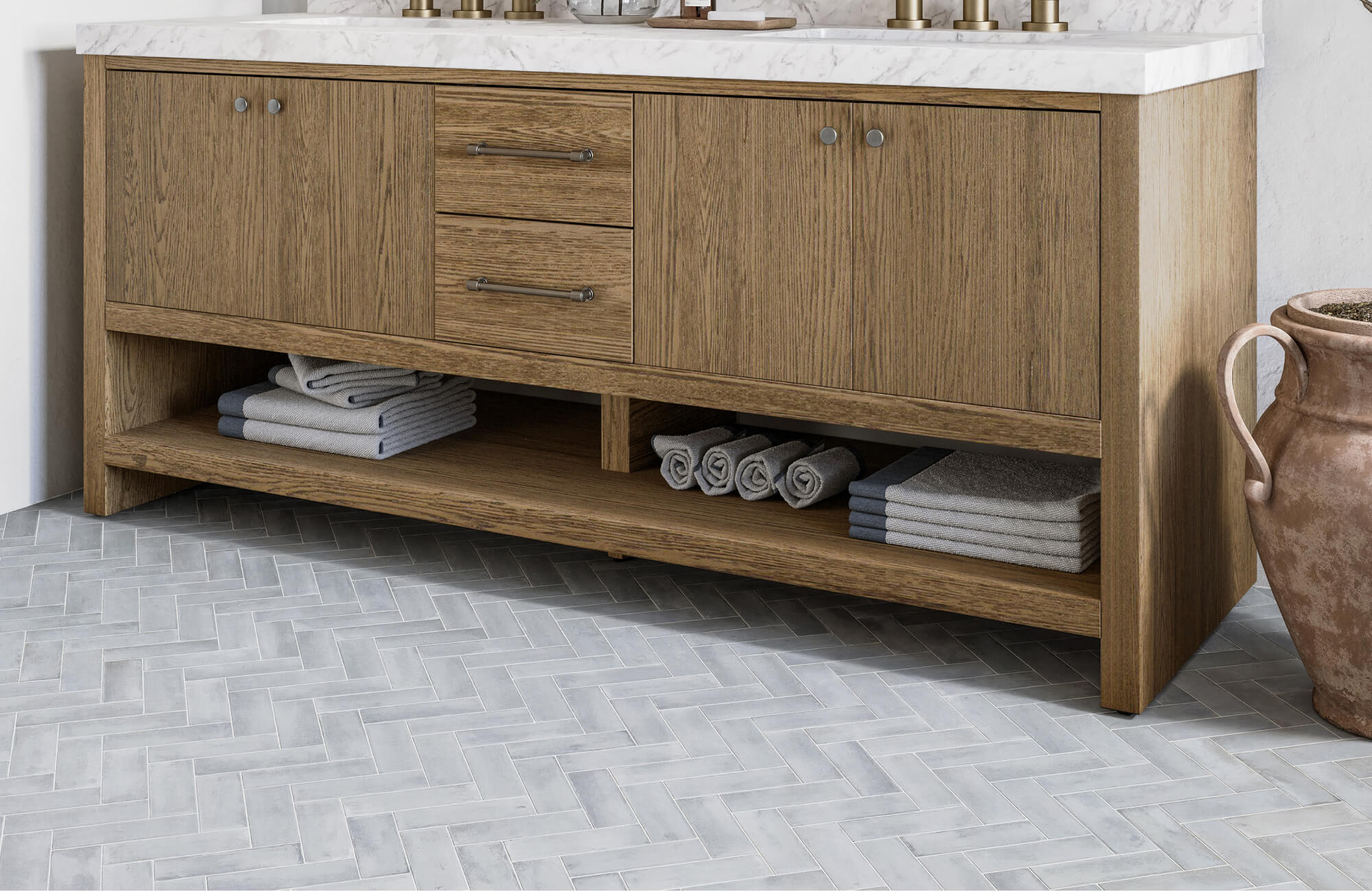 Gray subway tile floor installed in a herringbone pattern beneath a wooden bathroom vanity with open shelving and folded towels.