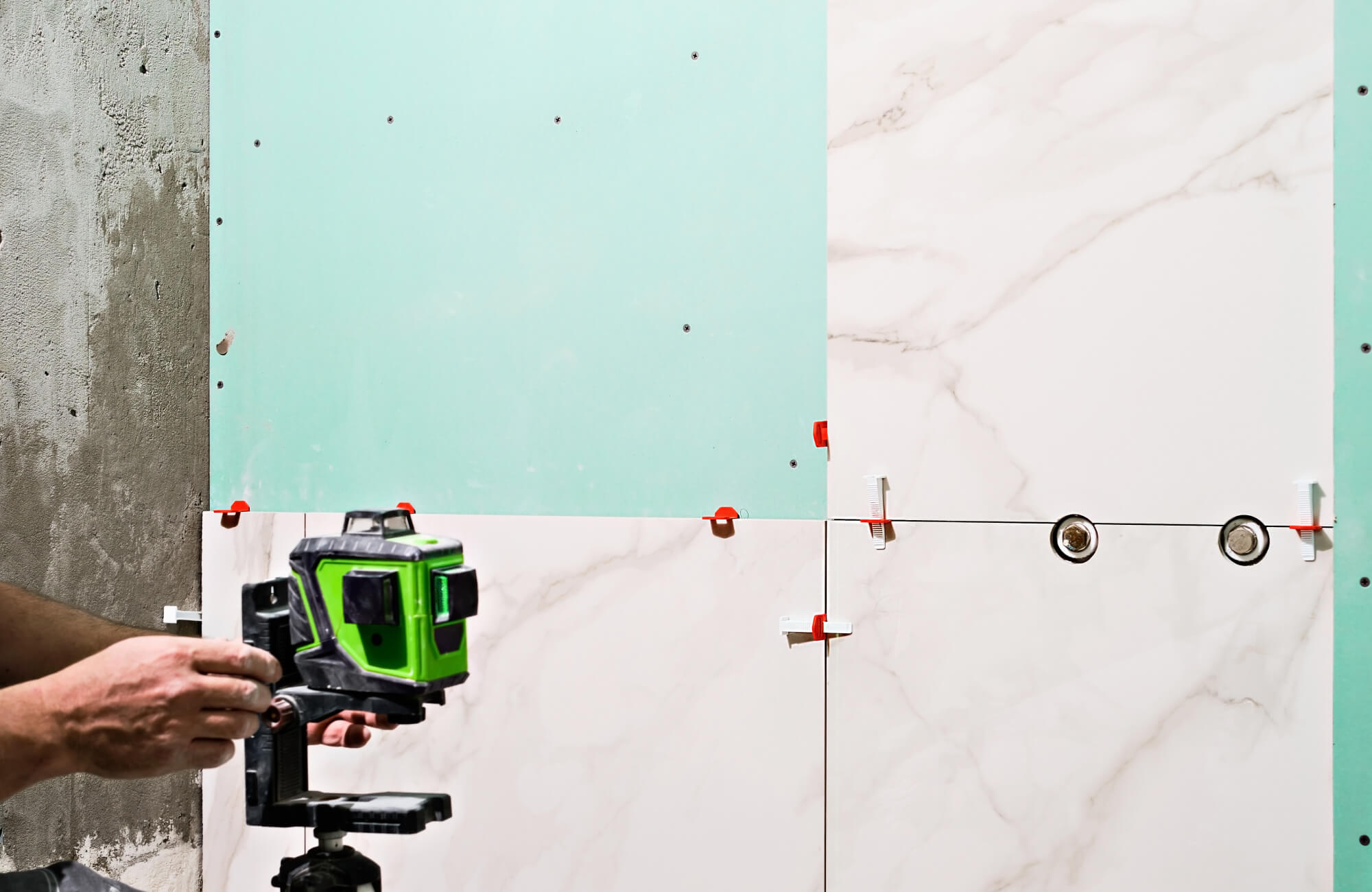 Large marble-look tiles installed on a green drywall bathroom wall using laser leveling and spacers.