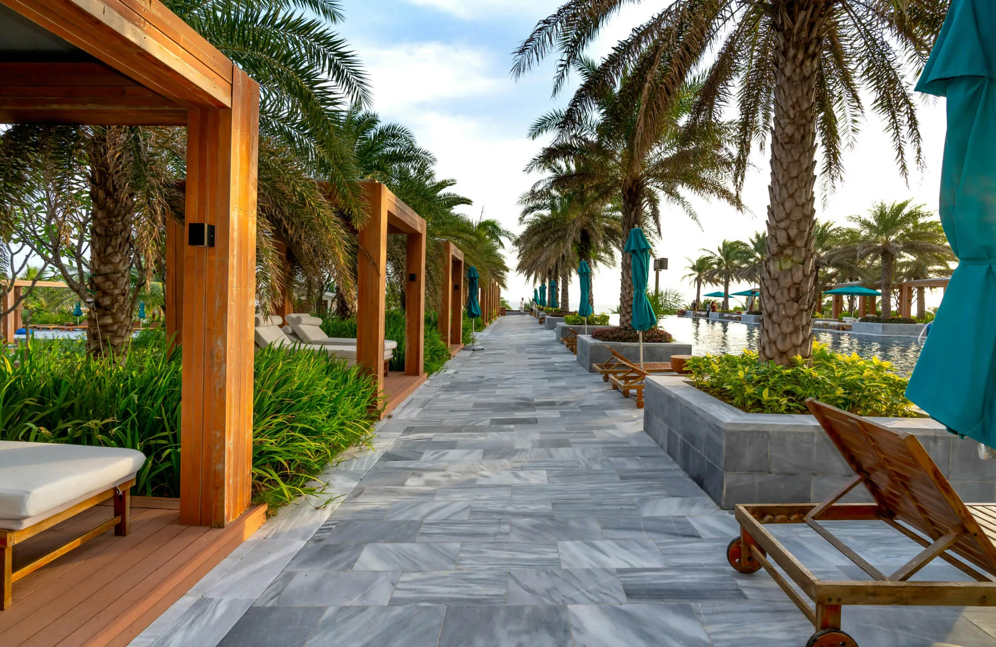 Marble-tiled walkway beside a resort pool with palm trees, lounge chairs, cabanas, and landscaped greenery.