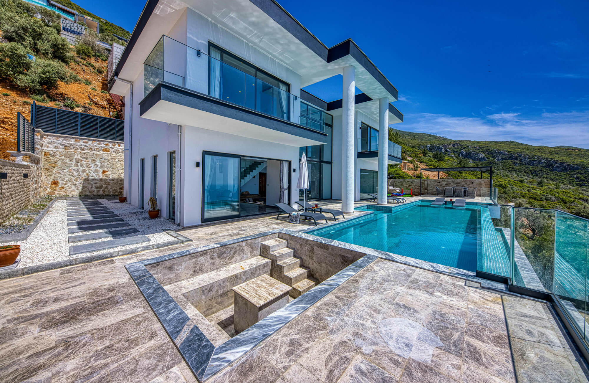 null Modern hillside home with marble-style outdoor flooring around a pool, sunken seating area, glass railings, and mountain views.